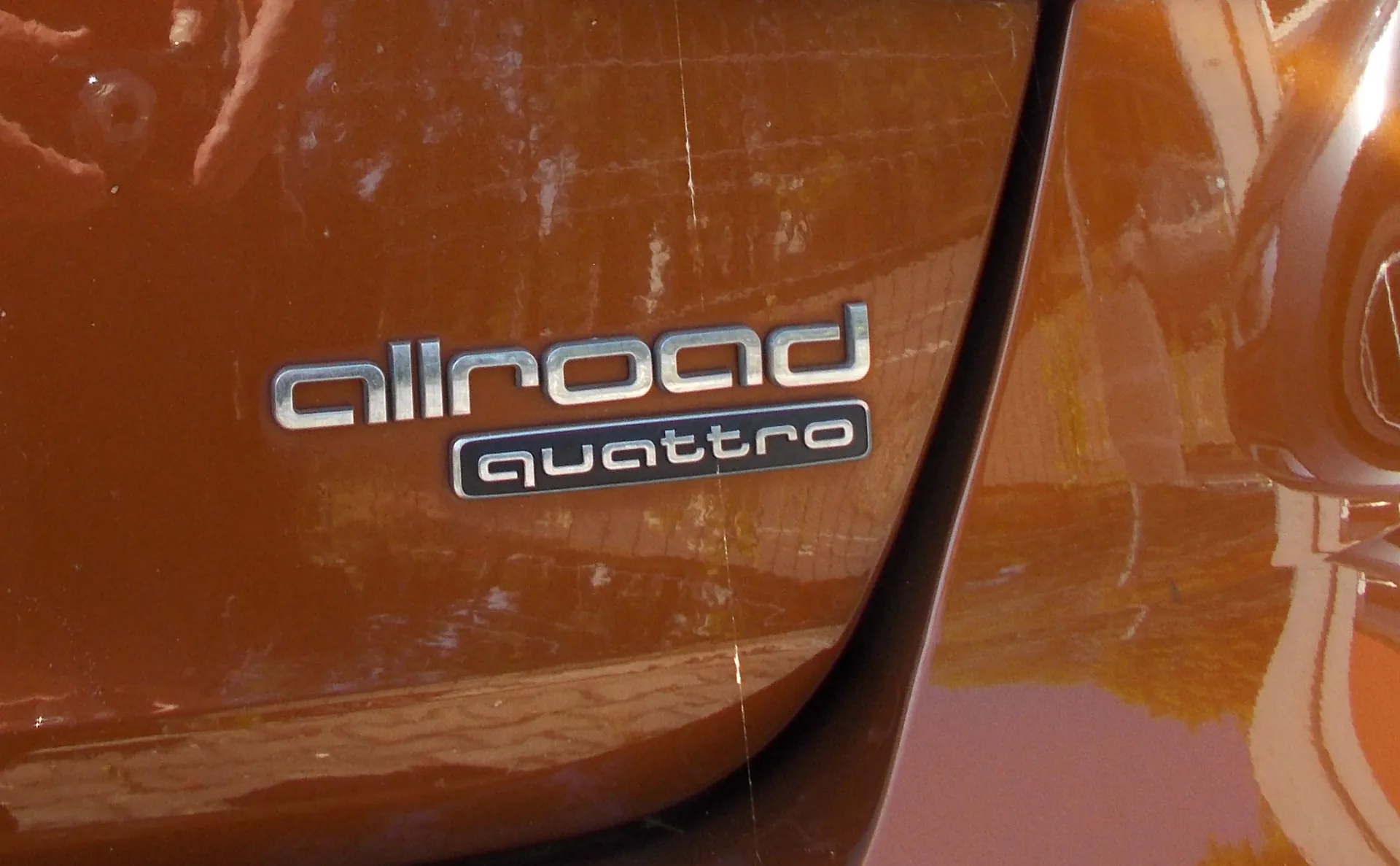 Zbliżenie na tylną część brązowego samochodu z chromowanymi emblematami. Na karoserii widnieje napis „allroad”, a poniżej mniejszy napis „quattro”. Powierzchnia lakieru jest błyszcząca i odbija otoczenie.