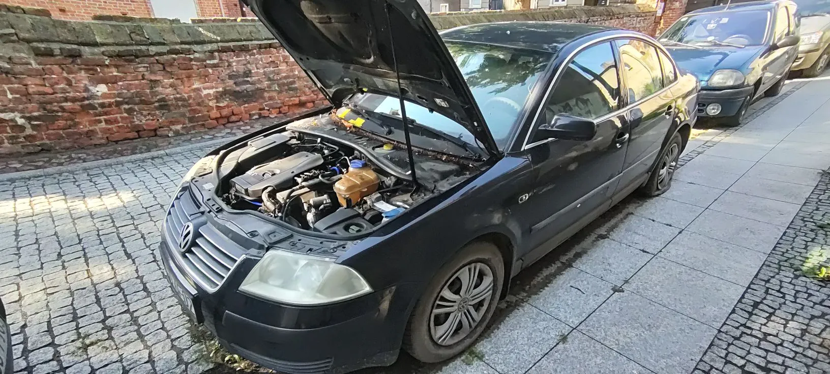 Czarny samochód osobowy marki Volkswagen zaparkowany na chodniku obok ceglanego muru. Maski samochodu jest otwarta, odsłaniając silnik i elementy komory silnika. W tle widoczny jest inny, ciemny samochód zaparkowany za nim.