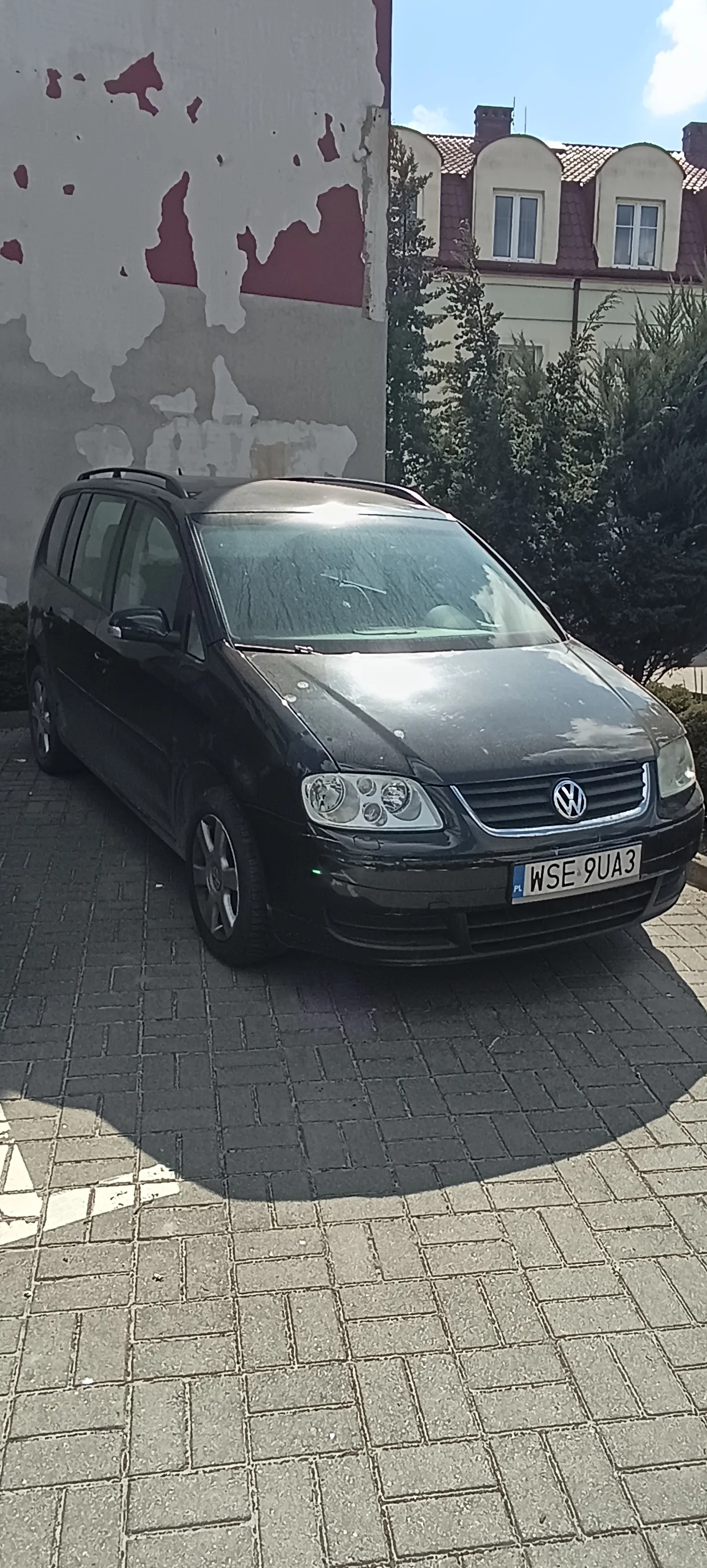 Czarny samochód Volkswagen zaparkowany na brukowanej powierzchni. Widoczna tablica rejestracyjna z napisem WSE 9UA3 oraz flagą PL. Po lewej stronie budynek z obłupioną farbą w kolorze szaro-czerwonym. W tle dom z czerwonym dachem, oknami dachowymi i zielonymi krzewami. Niebo jest jasne z kilkoma chmurami.