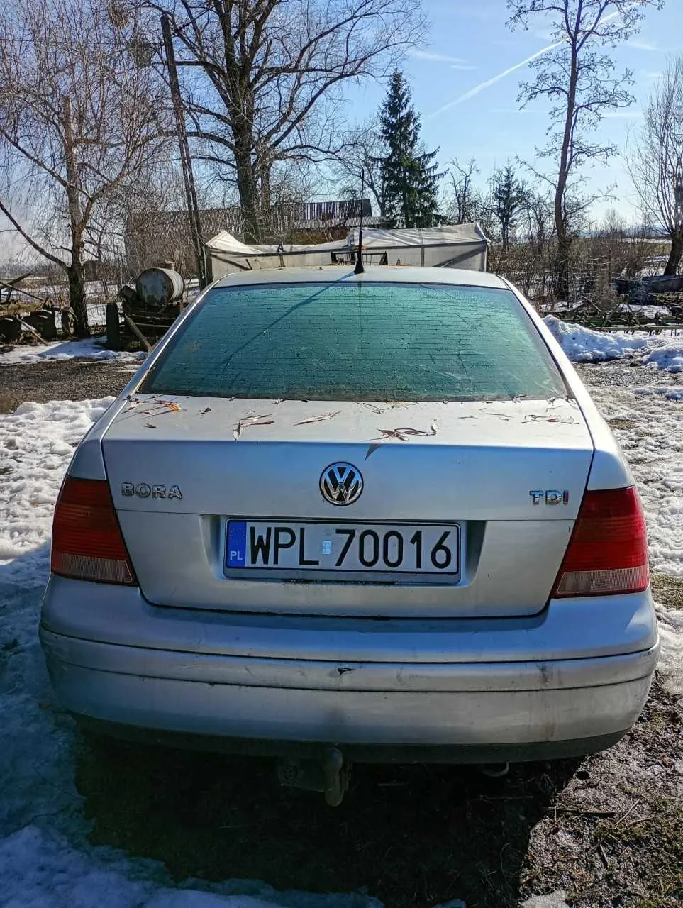 Tył srebrnego samochodu Volkswagen Bora TDI z tablicą rejestracyjną WPL 70016 (PL). Samochód stoi na podwórku z resztkami śniegu, w tle suche drzewa, namiot i budynki. Na bagażniku widoczne są liście, a na niebie biegną chmury. Oznaczenia 'BORA' i 'TDI' umieszczone są po bokach emblematu VW.