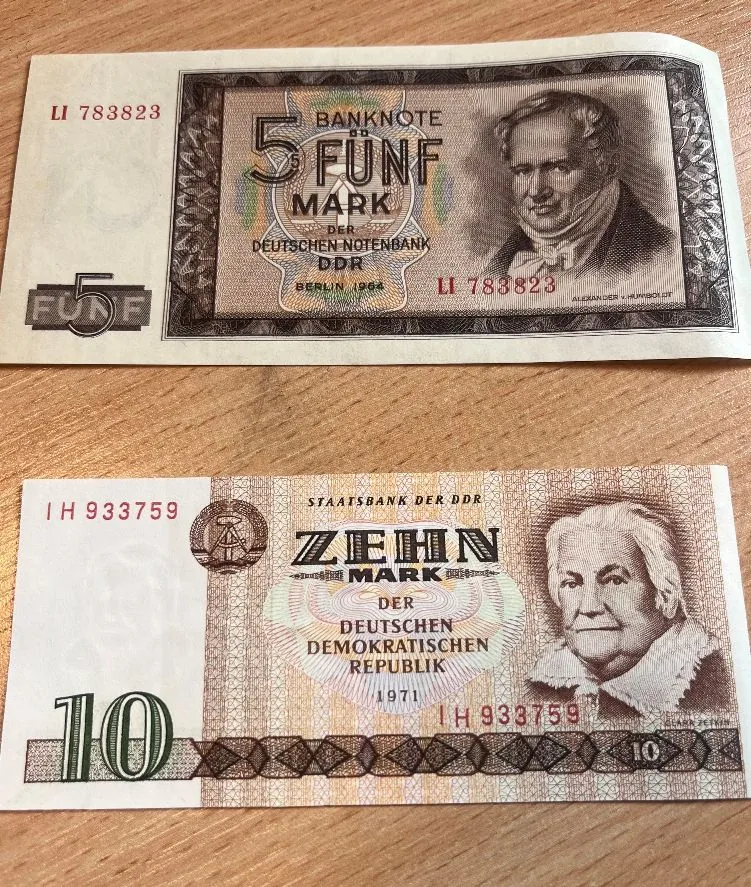 Dwa banknoty z Niemieckiej Republiki Demokratycznej (NRD) ułożone pionowo jeden nad drugim na drewnianym blacie. Górny banknot ma nominał 5 Mark (Fünf Mark) z wizerunkiem mężczyzny w profilu i numerem seryjnym LI 783823. Dolny banknot ma nominał 10 Mark (Zehn Mark) z wizerunkiem starszej osoby i numerem seryjnym IH 933759.