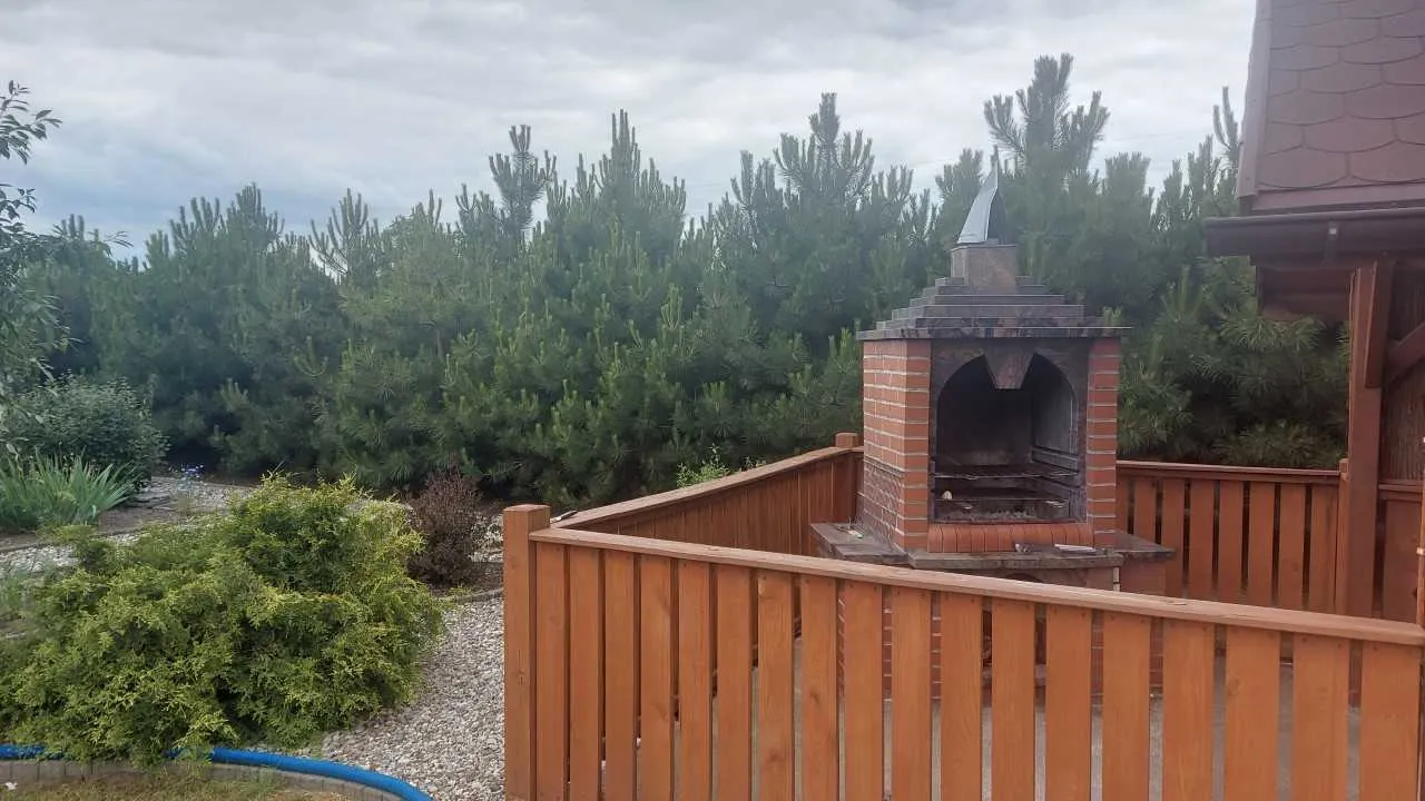 Zewnętrzny widok z ceglanym grillem z kominkiem na drewnianym tarasie z balustradą. Po prawej stronie widoczna część domu z brązowym dachem gontowym. W tle wysokie zielone drzewa iglaste, krzewy oraz ścieżka z kamieni. Niebo zachmurzone.