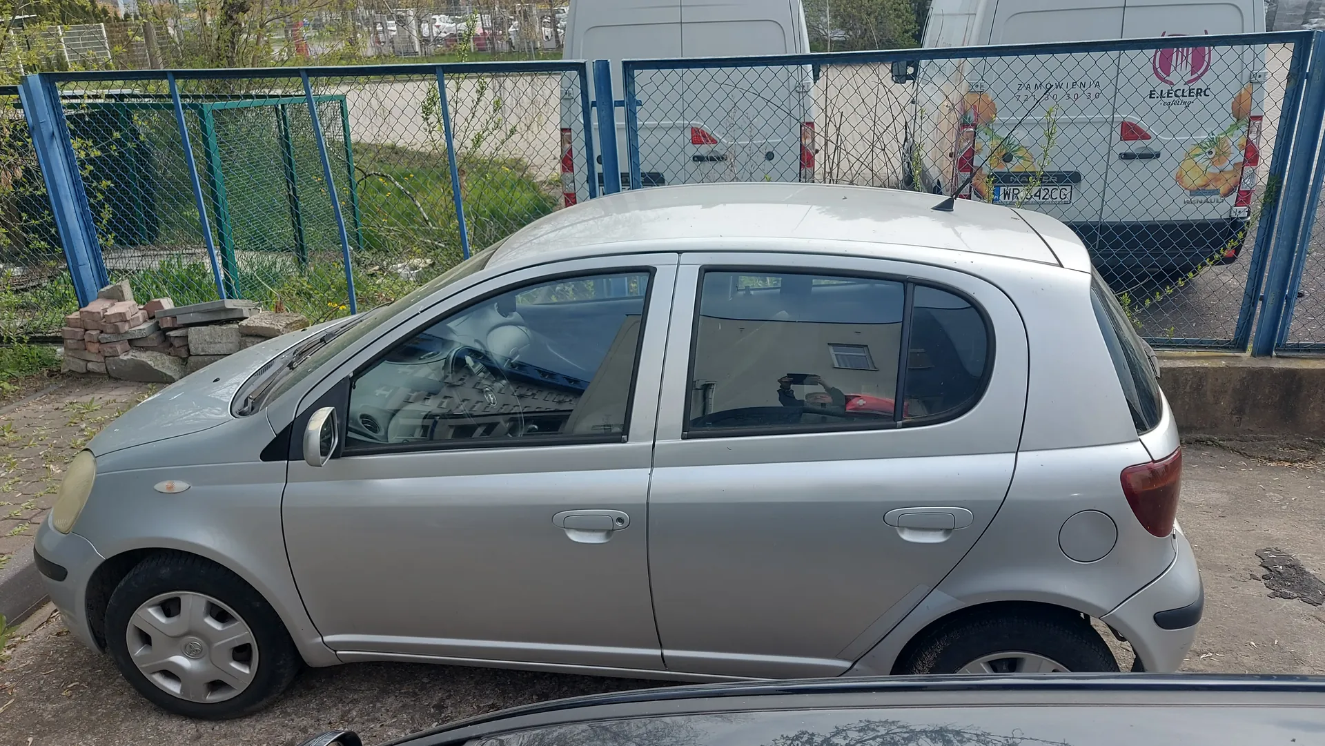 Srebrny samochód osobowy typu hatchback zaparkowany przed siatkowym ogrodzeniem z niebieskimi słupkami. W tle dwa białe furgony, z których jeden ma widoczny napis E.LECLERC, ZAMÓWIENIA oraz numer rejestracyjny WR 642CG. Po lewej stronie ogrodzenia stos cegieł i zieleń. Na pierwszym planie fragment czarnego pojazdu. Tekst na furgonie jest czytelny, horyzont jest poziomy.
