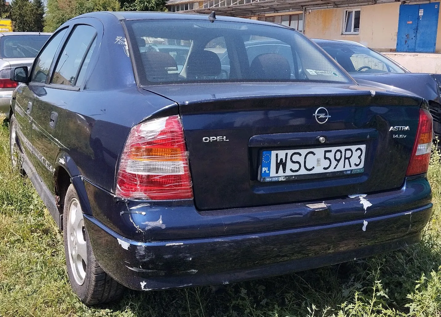 Tylny widok granatowego samochodu marki Opel Astra. Pojazd posiada polską tablicę rejestracyjną o numerze WSC 59R3. Na klapie bagażnika widoczne są napisy 'OPEL' oraz 'ASTRA 1.4 SE'. Lakier na tylnym zderzaku i boku nad kołem jest uszkodzony, z widocznymi odpryskami i zarysowaniami. Samochód stoi na trawie przed żółtym budynkiem.