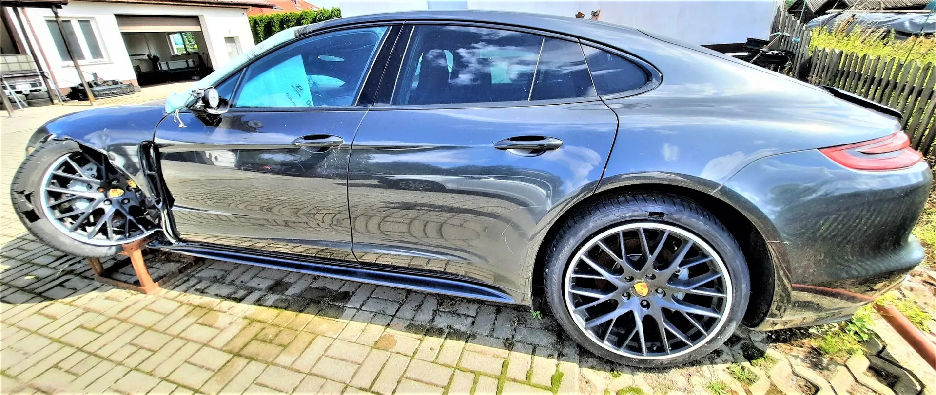 Zdjęcie przedstawiające ciemnoszary samochód marki Porsche Panamera z boku. Pojazd jest poważnie uszkodzony w przedniej części po stronie kierowcy – przednie lewe koło jest nienaturalnie wygięte i podparte drewnianym klockiem, a błotnik jest wgnieciony. Samochód stoi na kostce brukowej przed białym budynkiem z garażem.