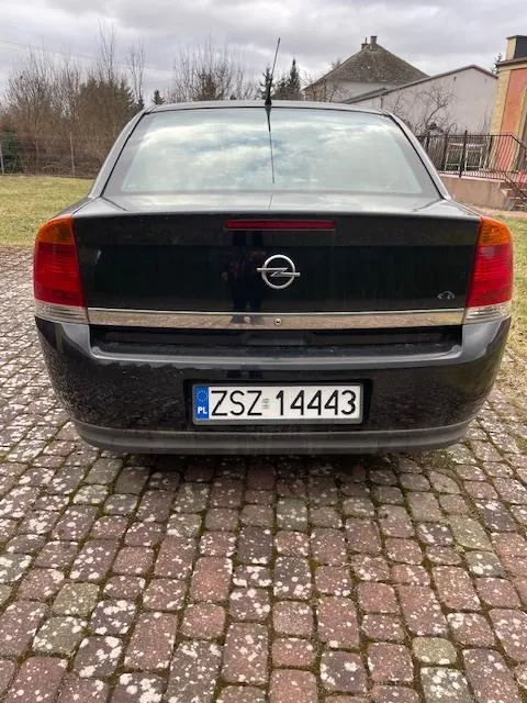 Tył czarnego samochodu marki Opel z tablicą rejestracyjną ZSZ 14443 z kodem PL po lewej stronie. Samochód stoi na brukowanej powierzchni. W tle widoczne są domy, drzewa bez liści oraz chmurne niebo. Czerwone światła tylnego oświetlenia i logo marki umieszczone są pośrodku bagażnika.