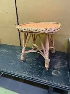 Widoczny jest stary, pleciony stołek z jasnym siedziskiem i drewnianymi nogami zawiązanymi sznurkiem. Obok stoi ciemnozielony stół z wyblakłą powierzchnią. Tło stanowi beżowa ściana z czarnym przewodem biegnącym poziomo. Całość ukazana jest w nieprawidłowej orientacji.