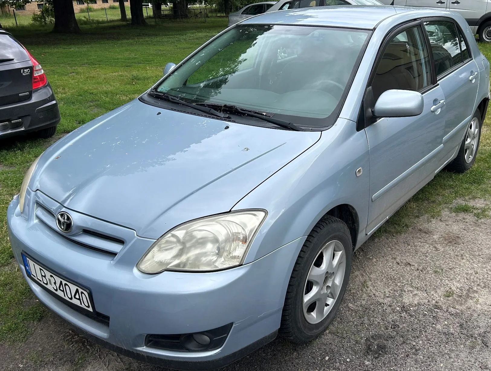 Jasnoniebieski samochód osobowy marki Toyota zaparkowany na żwirowym podłożu obok trawnika. Widoczny jest przód i prawy bok auta. Samochód posiada tablicę rejestracyjną o numerze LB 34040.