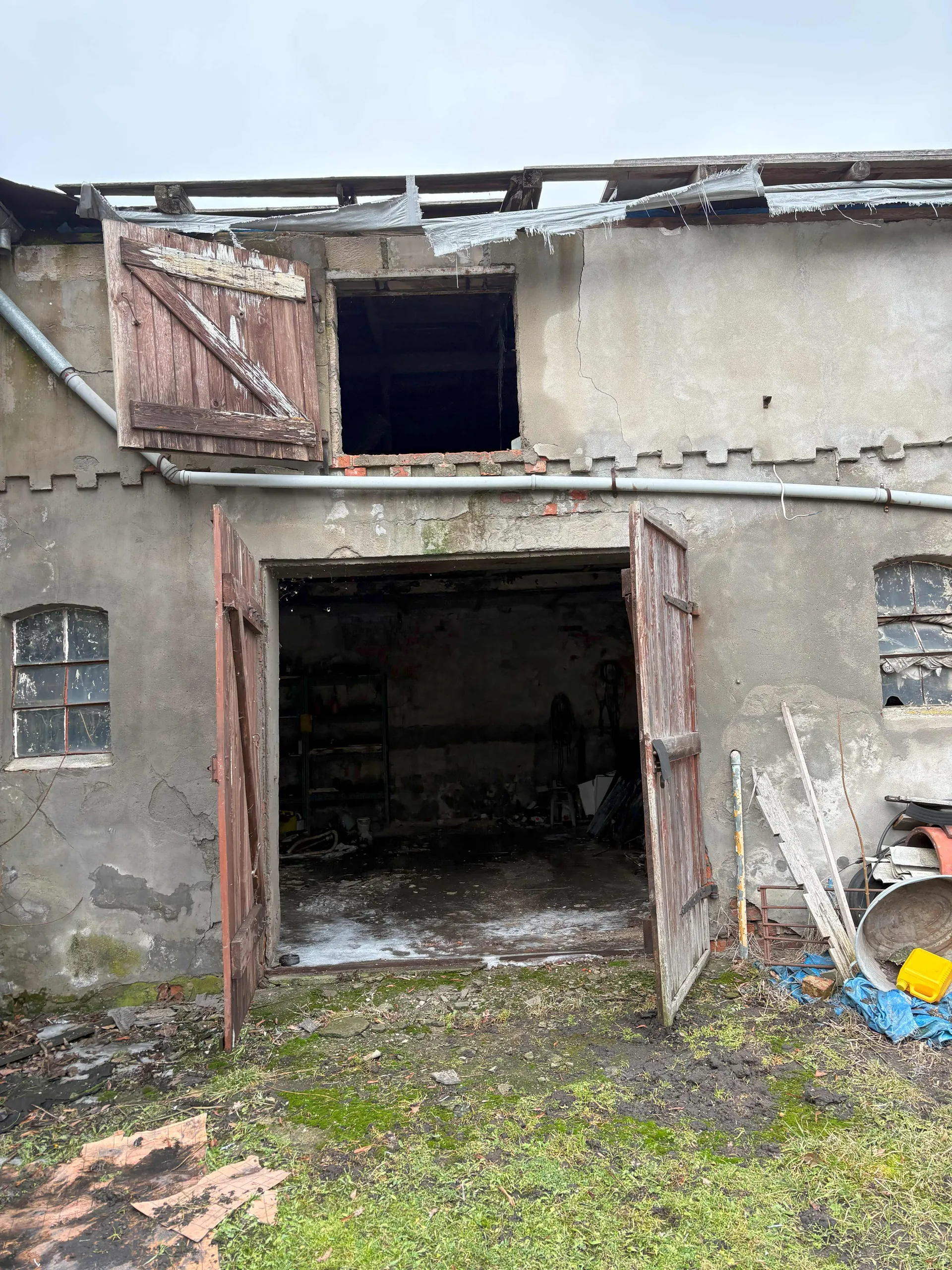 Zdjęcie przedstawia zaniedbaną, stary budynek o betonowych ścianach z widocznymi pęknięciami i plamami. W centrum znajdują się otwarte, drewniane drzwi prowadzące do ciemnego wnętrza z widocznymi półkami i narzędziami. Nad drzwiami otwarte okno z uszkodzonymi elementami konstrukcyjnymi oraz częściowo rozerwaną folią na dachu. Po lewej stronie małe, zniszczone okno z podzielonymi szybami, po prawej rozrzucone przedmioty: żółty pojemnik, niebieska folia, metalowe elementy i deski. Na ziemi trawa, mchy i gruz. Niebo jest pochmurne.