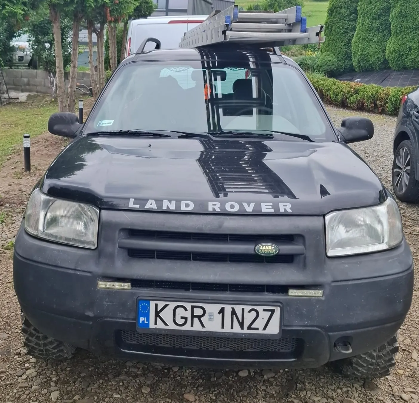 Czarny samochód marki Land Rover widziany od przodu. Na dachu pojazdu zamontowany jest bagażnik z aluminiową drabiną. Na masce widnieje napis 'LAND ROVER', a na grillu znajduje się małe logo marki. Samochód ma polską tablicę rejestracyjną o numerze KGR 1N27. Pojazd stoi na żwirowym podłożu, w tle widoczne są zielone krzewy i fragment białego vana.