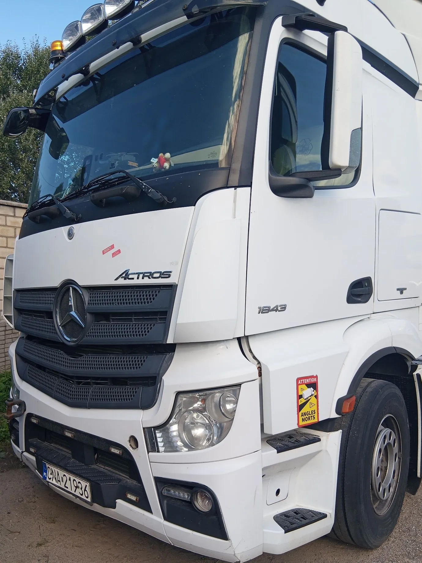 Zbliżenie na przód białej ciężarówki marki Mercedes-Benz Actros. Na grillu widoczne jest duże logo Mercedesy oraz napis 'Actros'. Po prawej stronie kabiny znajduje się oznaczenie modelu '1843'. Na błotniku przyklejona jest żółto-czerwona naklejka ostrzegawcza z napisem 'ATTENTION ANGLES MORTS'. Pojazd posiada tablicę rejestracyjną o numerze 'CNA 21936'. Na dachu zamontowane są dodatkowe reflektory i pomarańczowa lampka ostrzegawcza.