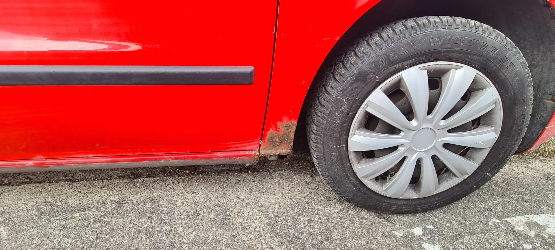 Zbliżenie na tylną część czerwonego samochodu, pokazujące koło z srebrną felgą aluminiową oraz fragment nadwozia z czarną listwą ochronną. Na dolnej krawędzi nadwozia, tuż przed nadkolem, widoczne są ślady korozji i rdzy.