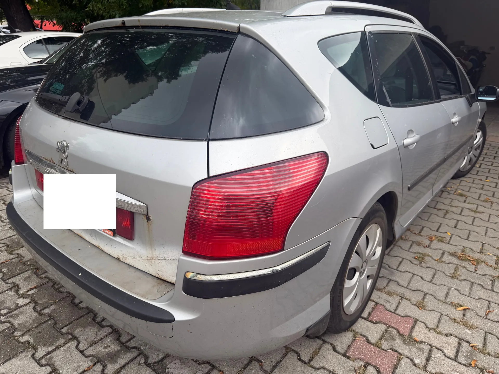 Srebrny samochód marki Peugeot w nadwoziu kombi, zaparkowany tyłem na kostce brukowej. Widoczny jest tylny zderzak, lampy oraz klapa bagażnika z logo producenta. Tablica rejestracyjna jest zakryta białym polem. Na lakierze w okolicach klapy bagażnika widoczne są ślady rdzy.