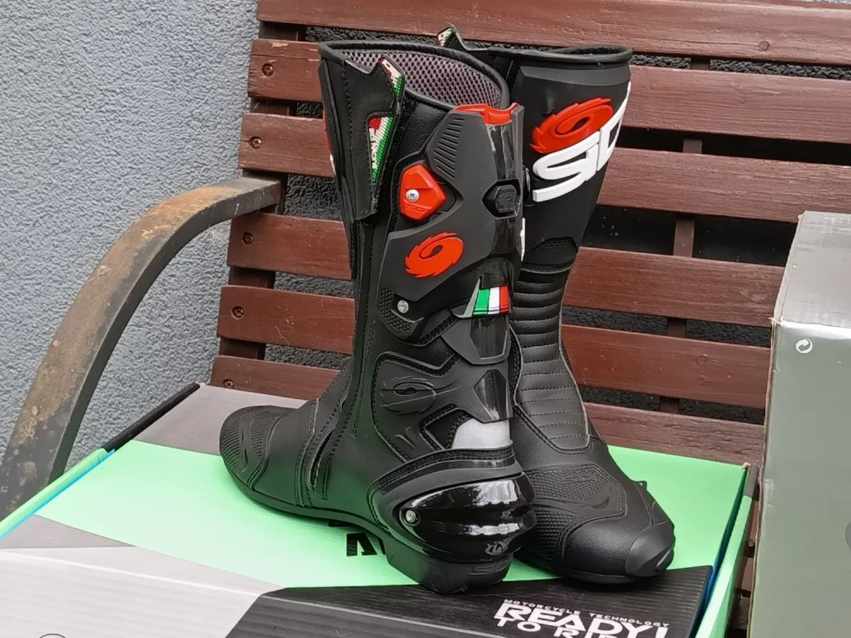 Para czarnych butów motocyklowych marki SIDI, ustawionych pionowo na zielono-białym pudełku, które znajduje się na brązowej drewnianej ławce. Buty mają czerwone i białe akcenty, w tym logo SIDI oraz małe flagi włoskie. Konstrukcja butów jest wzmocniona plastikowymi elementami ochronnymi.