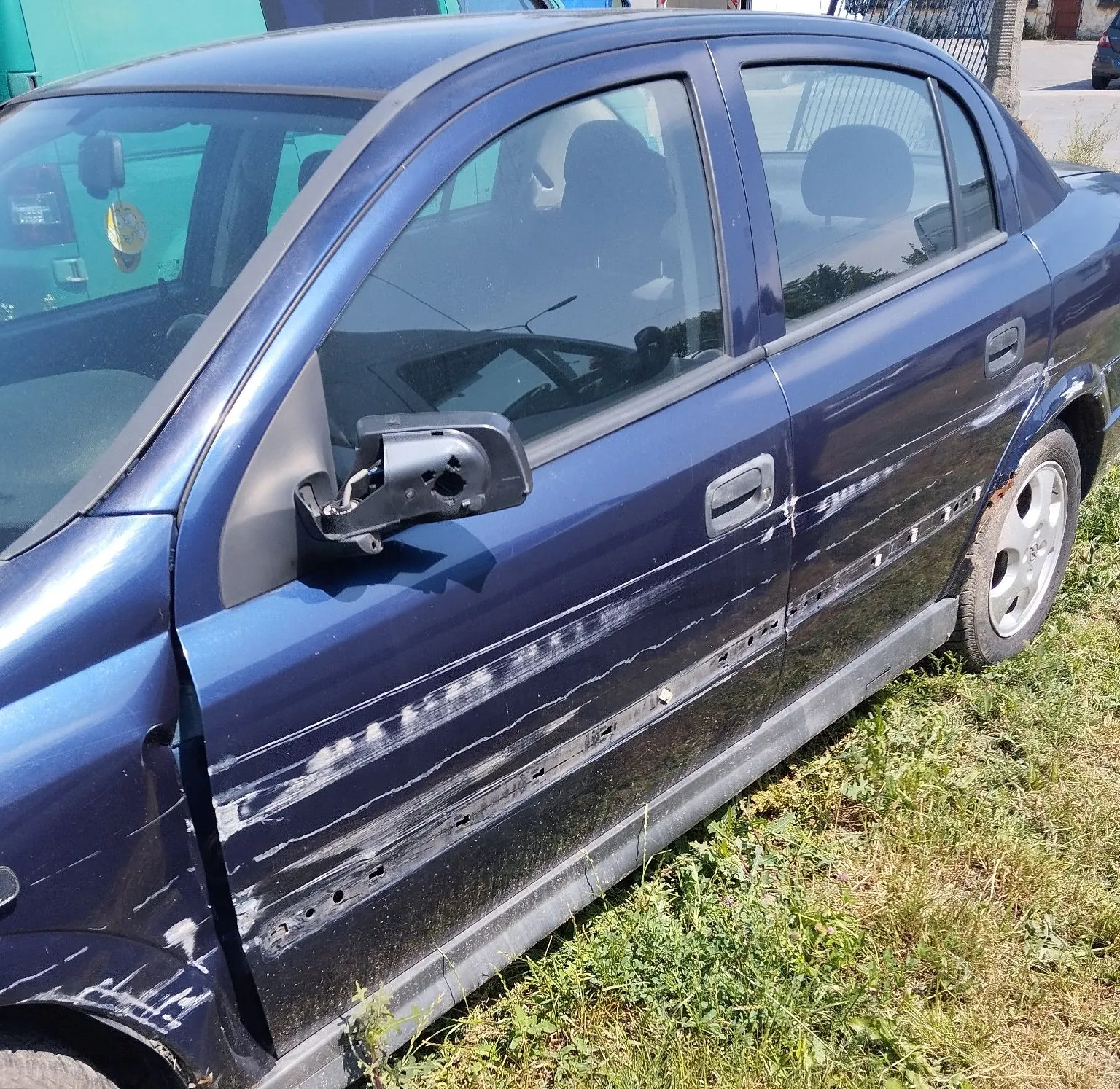 Zbliżenie na prawy bok ciemnoniebieskiego samochodu osobowego. Pojazd posiada liczne uszkodzenia: głębokie zarysowania i przetarcia lakieru wzdłuż przednich i tylnych drzwi oraz błotnika. Lusterko boczne jest uszkodzone i częściowo oderwane. Samochód stoi na trawiastym podłożu.