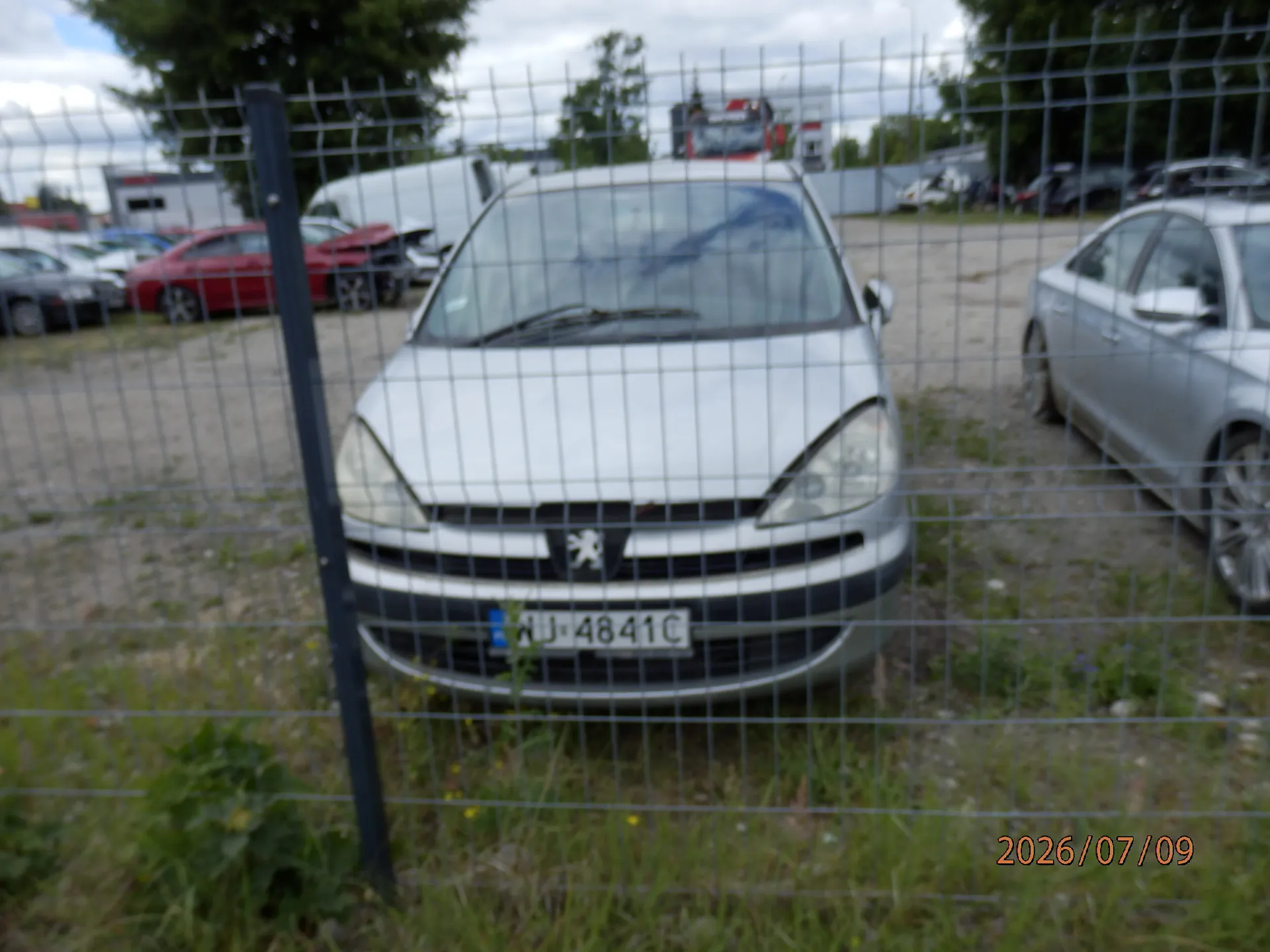 Srebrny samochód marki Peugeot zaparkowany na placu za metalowym ogrodzeniem z siatki. Na samochodzie widoczna jest tablica rejestracyjna o numerze AJ 4841C. W tle widoczne są inne zaparkowane pojazdy, w tym czerwony samochód oraz biały furgon. W prawym dolnym rogu obrazu znajduje się data 2026/07/09.
