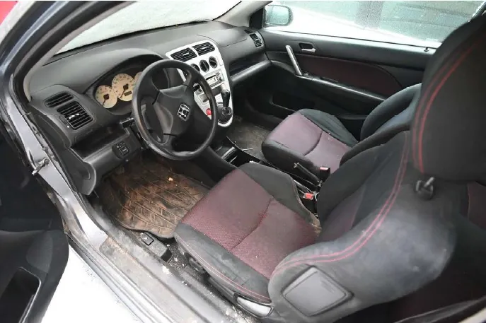 Wnętrze samochodu z otwartą lewą przodem drzwiami. Widoczna kierownica z logo Hondy, czarne siedzenia z czerwonym przeszyciem, zniszczone dywaniki na podłodze, deska rozdzielcza z dwoma okrągłymi wskaźnikami oraz centralna konsoła z dźwignią zmiany biegów. Wnętrze wydaje się nieczyste, z widocznymi śladami brudu na podłodze i siedzeniach.