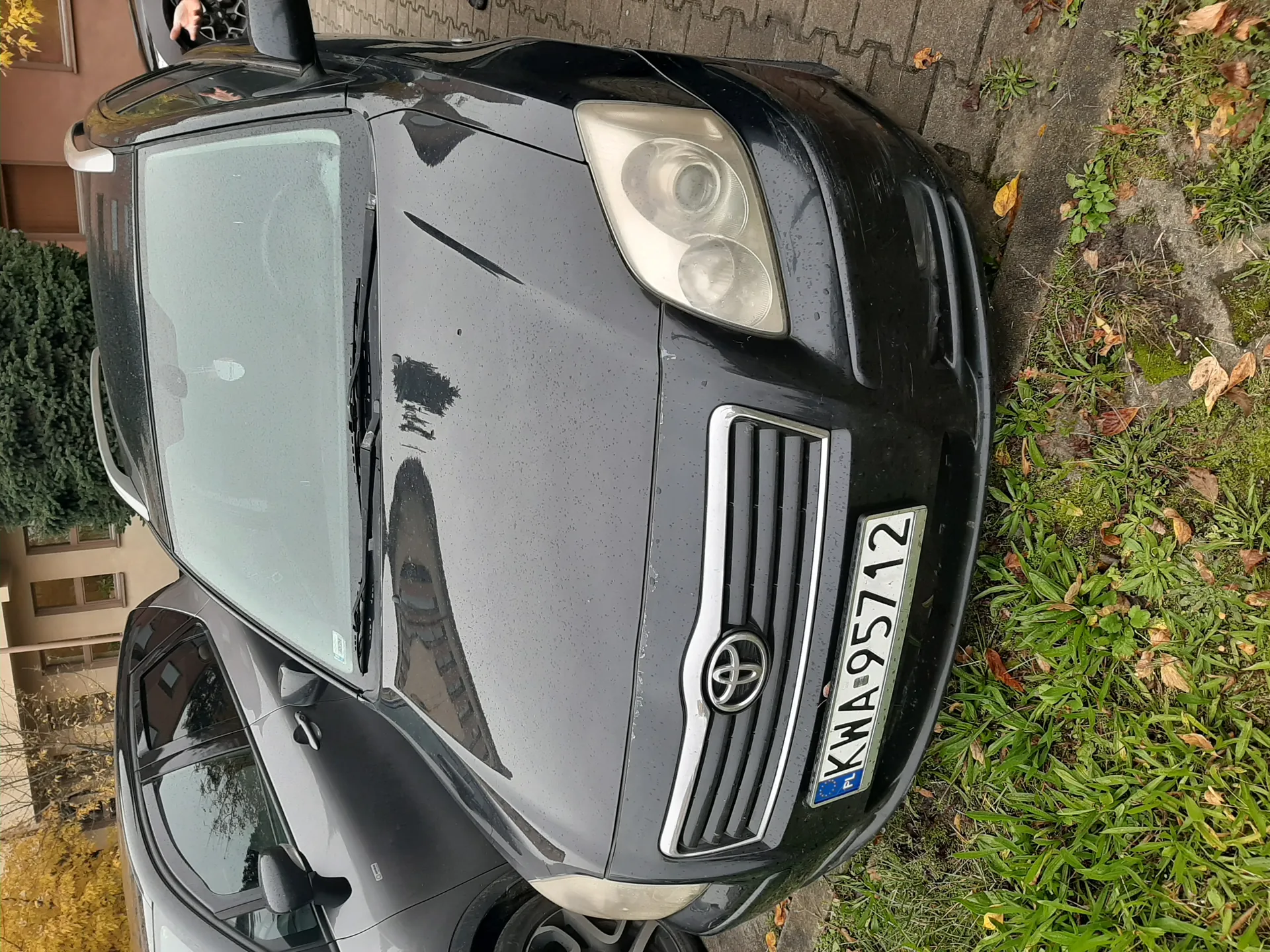 Czarny samochód marki Toyota z tablicą rejestracyjną KWA-95712 (PL) zaparkowany na brukowanej powierzchni obok trawnika z suchymi liśćmi. W tle widoczne są inne pojazdy, budynki mieszkalne oraz drzewa. Na przedniej szybie samochodu znajduje się drobna zawieszka.