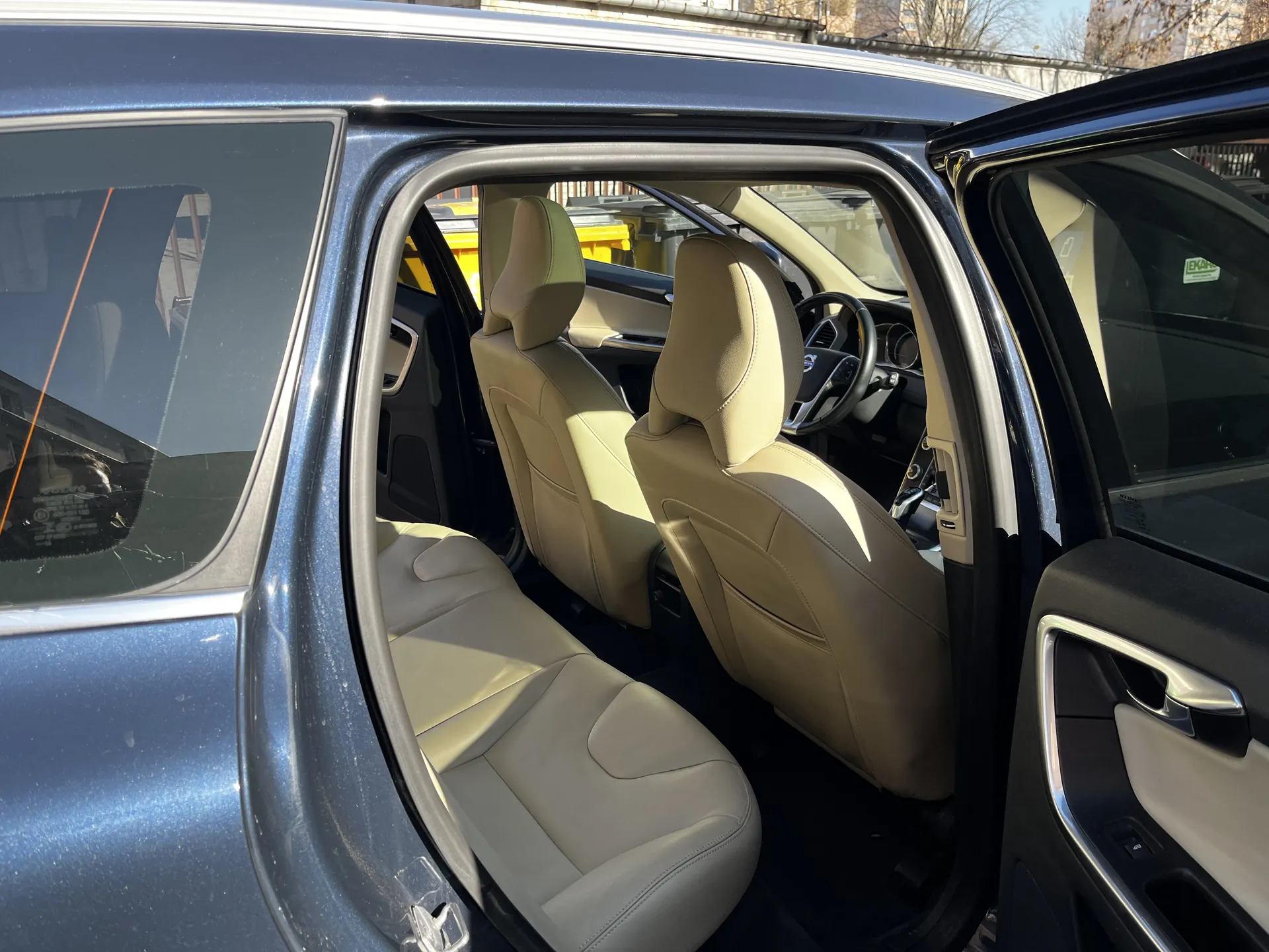 Wnętrze niebieskiego samochodu z otwartą drzwiami kierowcy. Widoczne beżowe skórzane fotele przodowe, kierownica, deska rozdzielcza oraz fragmenty zewnętrzne z żółtymi pojemnikami i budynkami na tle.