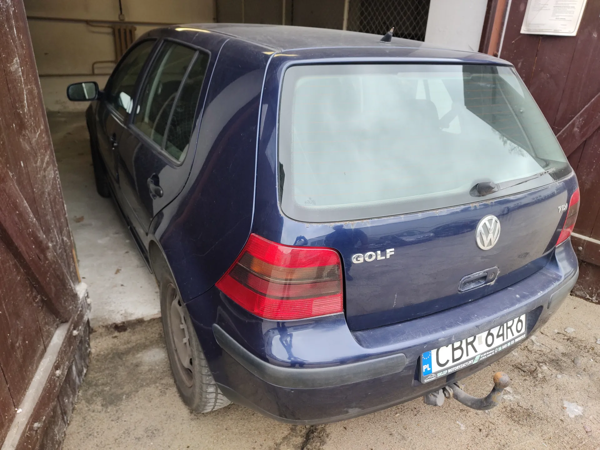 Ciemnoniebieski samochód osobowy Volkswagen Golf z tyłu, zaparkowany w garażu z brązowymi drzwiami po bokach. Na bagażniku widoczny napis GOLF oraz logo VW. Tablica rejestracyjna PL CBR 64R6 z napisem SKLEP MOTORYZACYJNY. Samochód ma czerwone światła tylna, hak holowniczy i lekko zabrudzoną karoserię. Podłoga garażu z betonu, a po prawej stronie widoczna tablica informacyjna na ścianie.