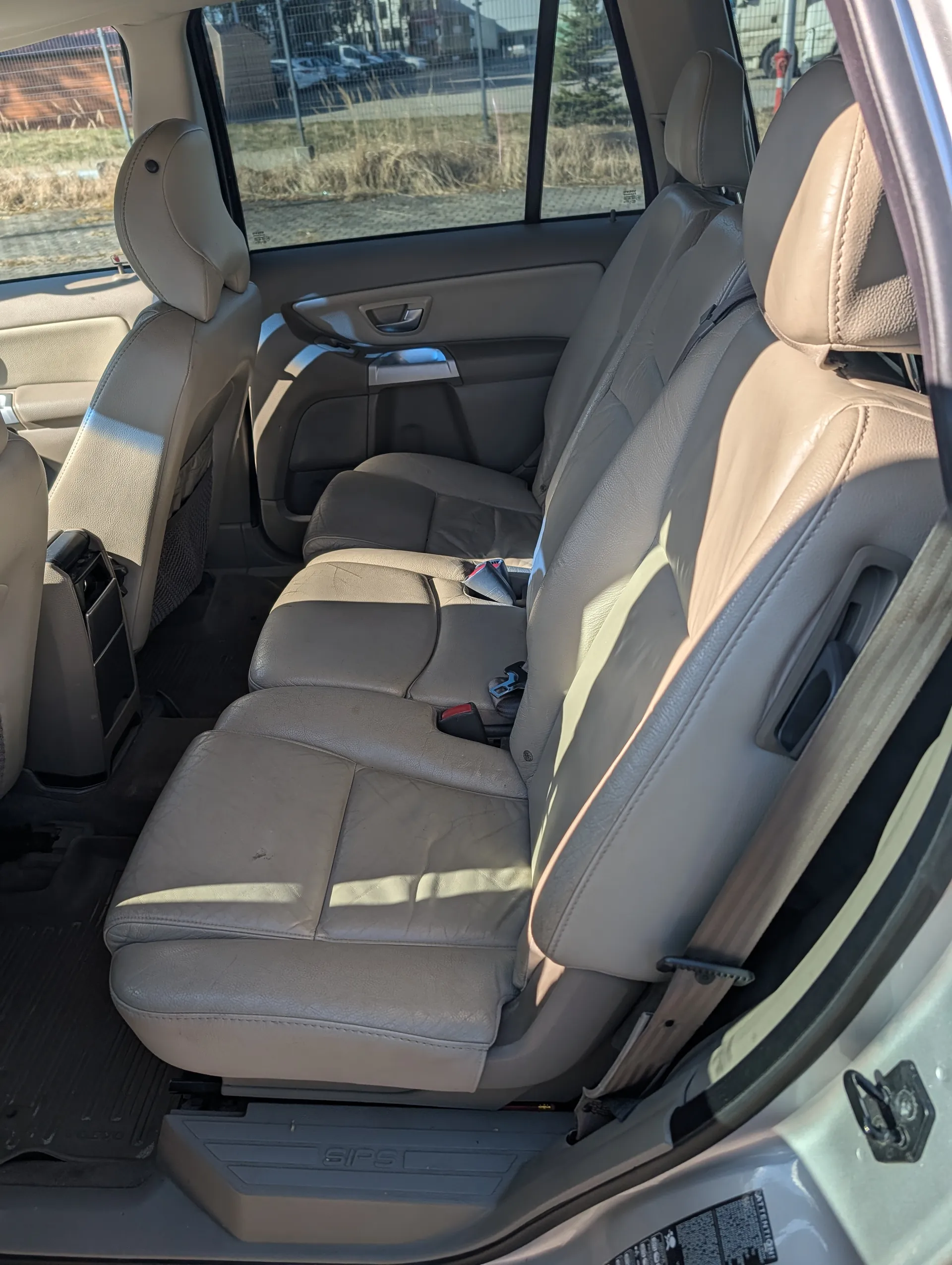 Wnętrze samochodu z beżowymi skórzonymi siedzeniami tylnymi. Widoczne są poduszki biodrowe, pasy bezpieczeństwa oraz otwarte drzwi po prawej stronie. Przez okna widać parking z innymi pojazdami i ogrodzenie. Na podłodze czarne maty, a na panelu bocznym napis 'SIPS'.