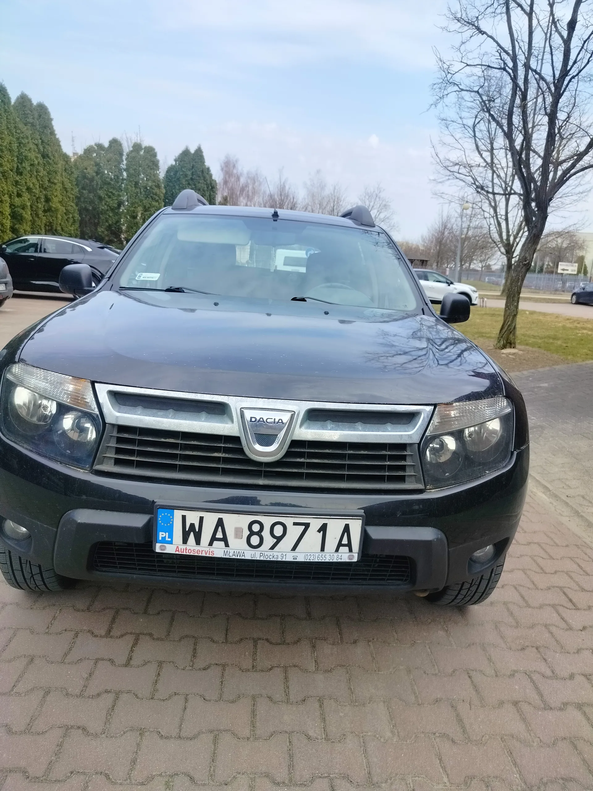 Czarny samochód marki Dacia z przodu, z widoczną tablicą rejestracyjną WA 8971A. Na tablicy widnieje napis 'PL', 'Autoservis', 'MŁAWA ul. Płocka 91' oraz numer telefonu '(023) 655 30 84'. W tle drzewa, inne zaparkowane samochody i niebo z chmurami. Samochód stoi na brukowanej powierzchni.