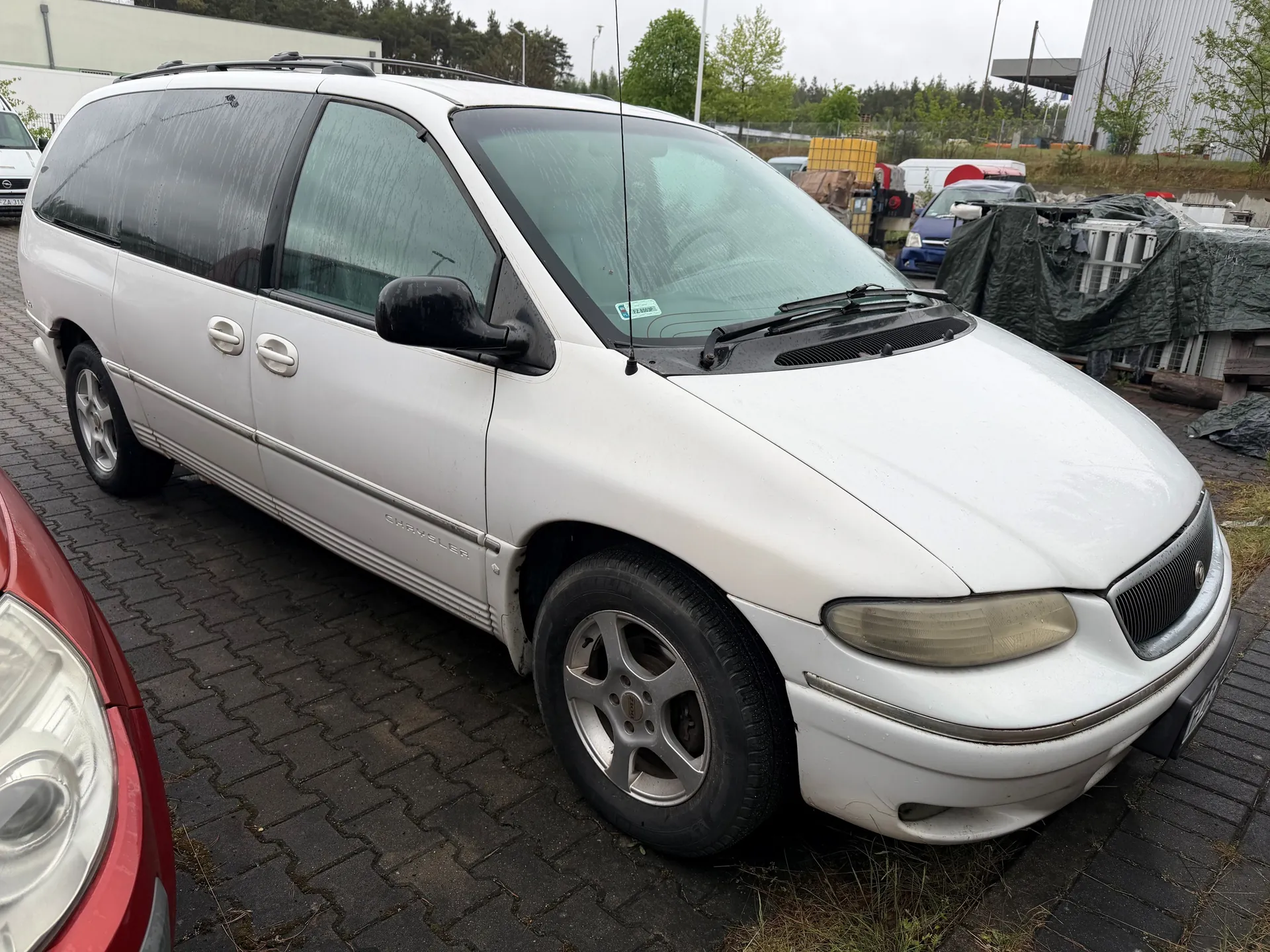 Biały minivan marki Chrysler zaparkowany na brukowanym placu. Po lewej stronie widoczna część czerwonego samochodu. W tle inne pojazdy, niektóre przykryte czarną folią, budynki przemysłowe, drzewa i pochmurne niebo. Na boku minivana widoczny napis 'Chrysler'.