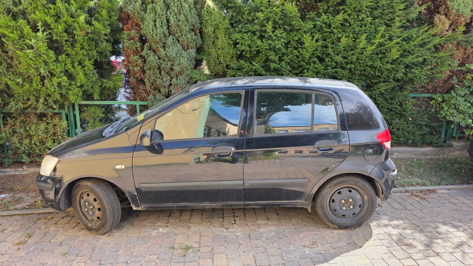 Czarny, pięciodrzwiowy samochód osobowy zaparkowany bokiem na kostce brukowej. W tle znajduje się gęsty, zielony żywopłot z krzewów oraz zielony metalowy płot.
