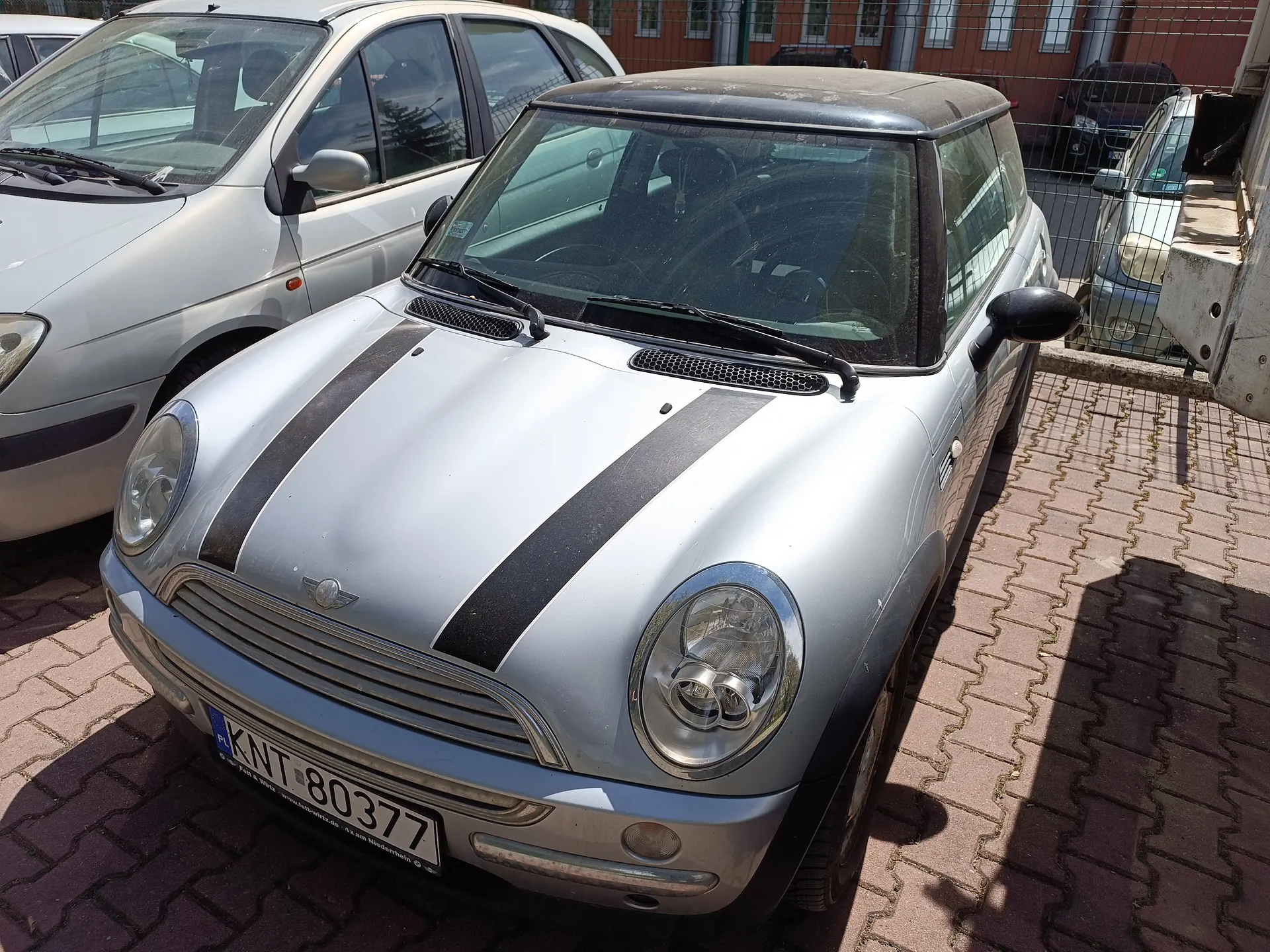 Srebrny samochód Mini Cooper z czarnymi paskami na masce, z tablicą rejestracyjną KNT 80377 (z lewej strony widoczny znak PL). Pojazd zaparkowany na brukowanej powierzchni, w tle inne samochody, metalowe ogrodzenie oraz czerwony budynek z oknami. Wnętrze samochodu i elementy takie jak reflektory, kierownica oraz deska rozdzielcza są widoczne przez przednią szybę.