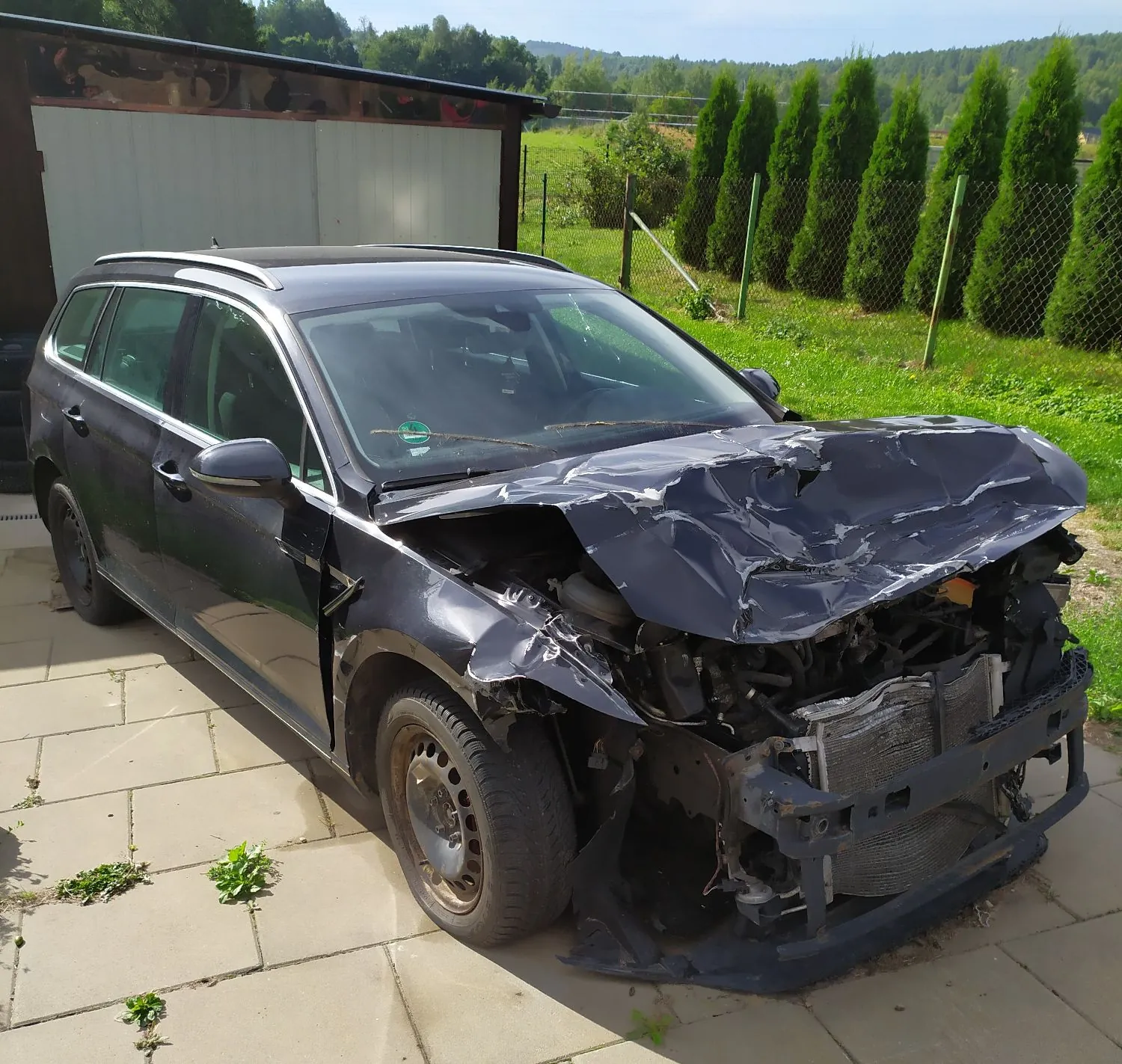 Czarny samochód typu kombi z poważnie uszkodzonym przodem. Maską jest mocno wgnieciona i pognieciona, zniszczony jest zderzak oraz widoczne są uszkodzenia chłodnicy i elementów konstrukcyjnych przodu pojazdu. Samochód stoi na betonowym podjeździe przed białym garażem, a w tle widać zielony trawnik i rząd wysokich, wąskich drzew iglastych.