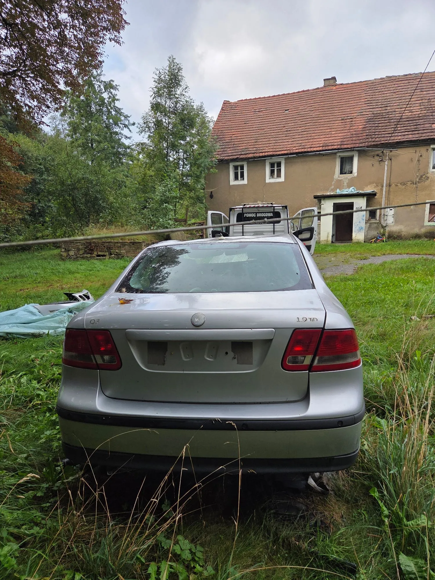 Srebrny samochód osobowy zaparkowany tyłem na trawiastym terenie. Pojazd nie posiada tablic rejestracyjnych, a na klapie bagażnika widnieje napis '1.9 TDI'. W tle znajduje się biały samochód dostawczy z napisem 'POMOC DROGOWA' oraz stary, jednopiętrowy dom z czerwonym dachem i szarymi ścianami.