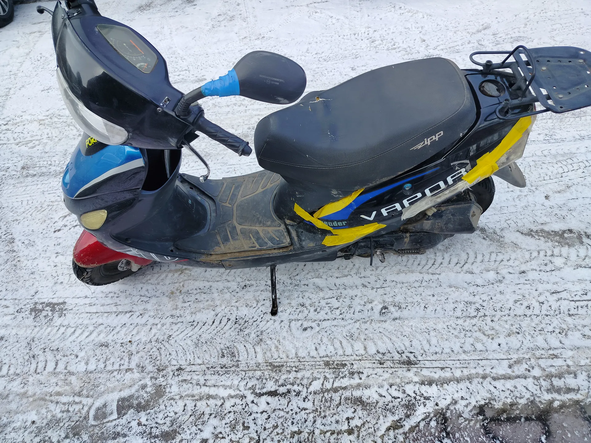Ciemnoniebieski skuter z niebieskimi i żółtymi akcentami, zaparkowany na śnieżnej nawierzchni z odciskami opon. Czarna siedzisko z napisem "zipp". Na boku skutera widoczny biały napis "VAPOR" oraz żółta taśma. Przednia część w kolorze niebieskim z białą listwą i żółtym napisem "FOR". Czerwona część koła przedniego. Niebieska rękojeść na jednym z kierownic. Tablica przyrządów z widocznym wskaźnikiem prędkości. Skuter oparty na podstawce.