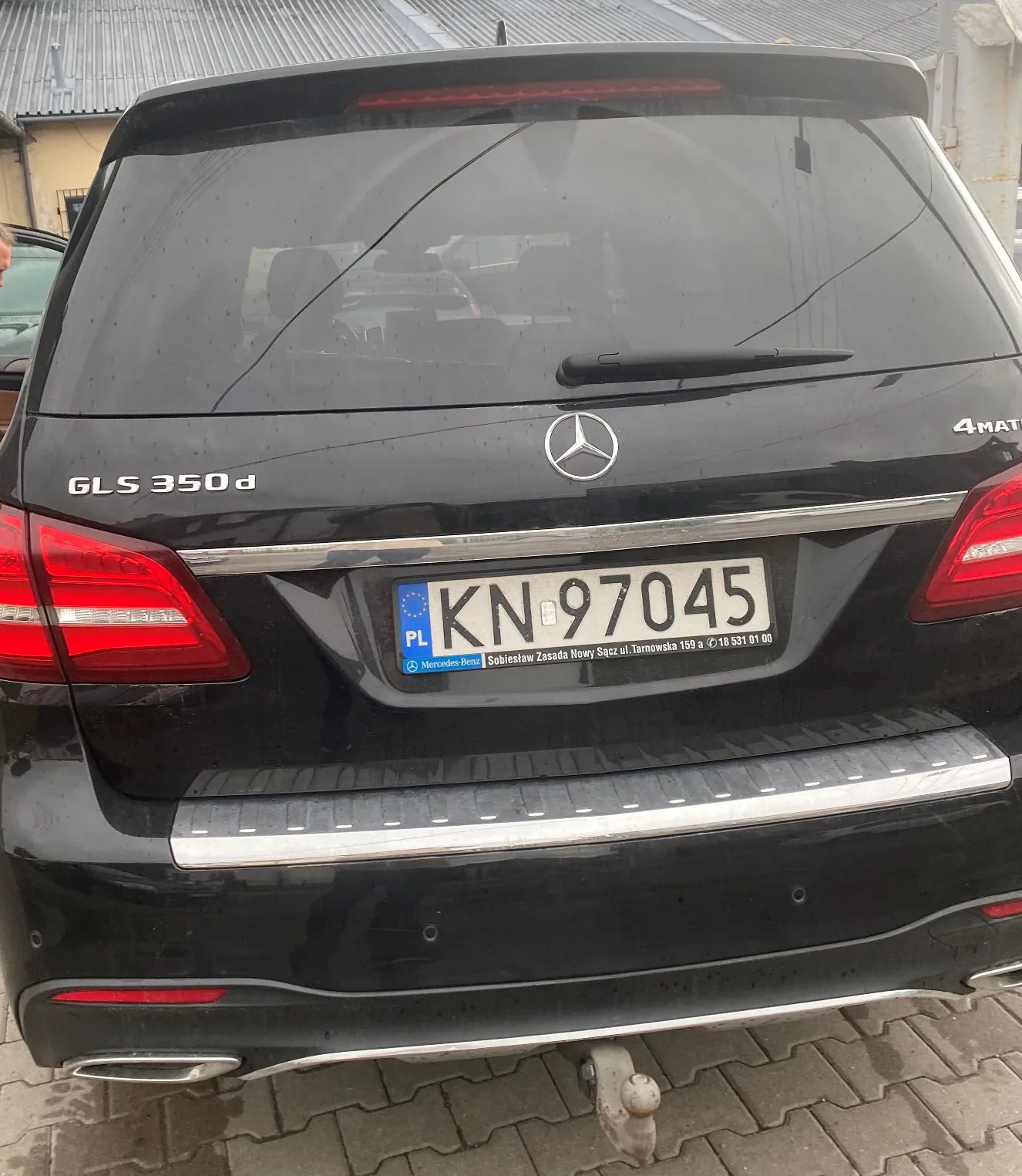 Tylny widok czarnego samochodu marki Mercedes-Benz. Na klapie bagażnika widoczne są oznaczenia 'GLS 350 d' po lewej stronie oraz '4MATIC' po prawej. W centrum znajduje się logo Mercedesa. Samochód posiada polską tablicę rejestracyjną o numerze 'KN 97045'. Poniżej tablicy znajduje się napis: 'Mercedes-Benz Sobiesław Zasada Nowy Sącz ul. Tarnowska 159 a ☎ 16 531 01 00'. Widoczne są czerwone tylne lampy oraz chromowane wykończenia zderzaka i klapy.