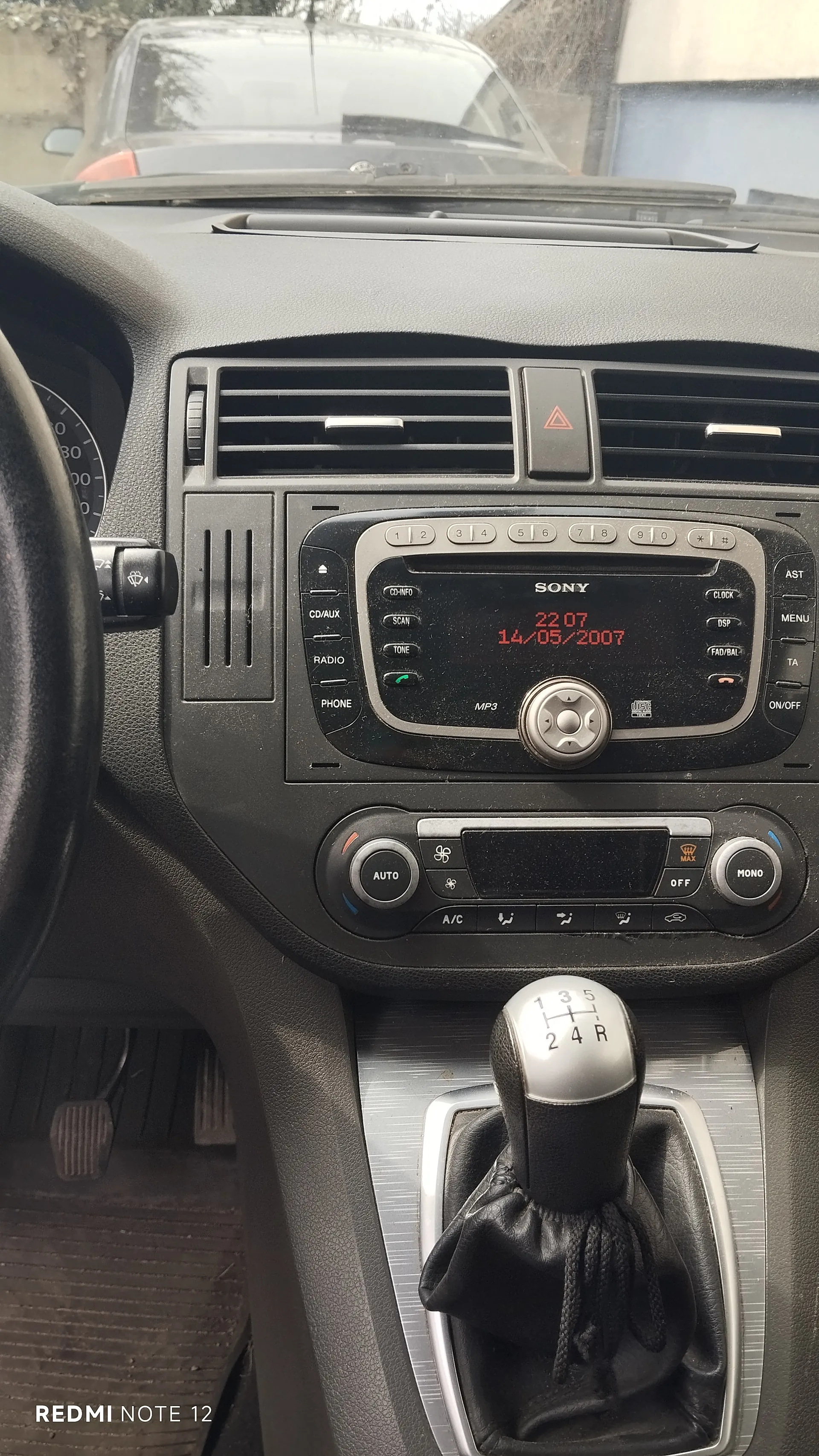 Wnętrze samochodu z widokiem na deski rozdzielczą. Po lewej stronie kierownica, w centrum radio Sony z wyświetlaczem pokazującym godzinę 22:07 i datę 14/05/2007. Poniżej radio znajduje się klimatyzacja z przyciskami AUTO, A/C oraz pokrętłami. W dolnej części widoczna dźwignia biegów z oznaczeniami 1, 2, 3, 4, 5, R. Przez przednią szybę widać inny samochód. W lewym dolnym rogu napis REDMI NOTE 12.