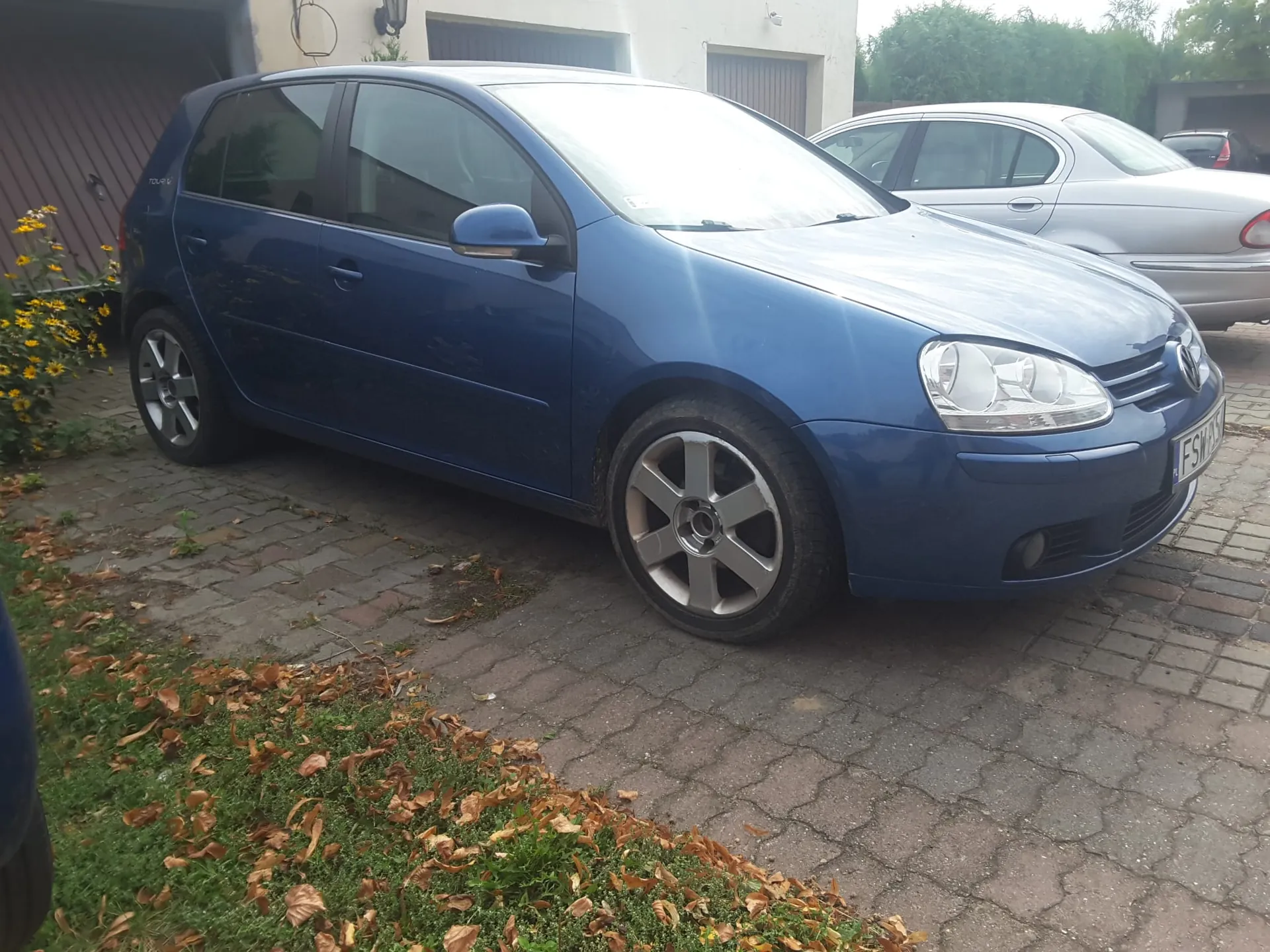 Niebieski samochód osobowy marki Volkswagen Golf zaparkowany na kostce brukowej przed budynkiem. Obok stoi srebrny samochód. Na pierwszym planie widoczny jest trawnik z opadłymi jesiennymi liśćmi.