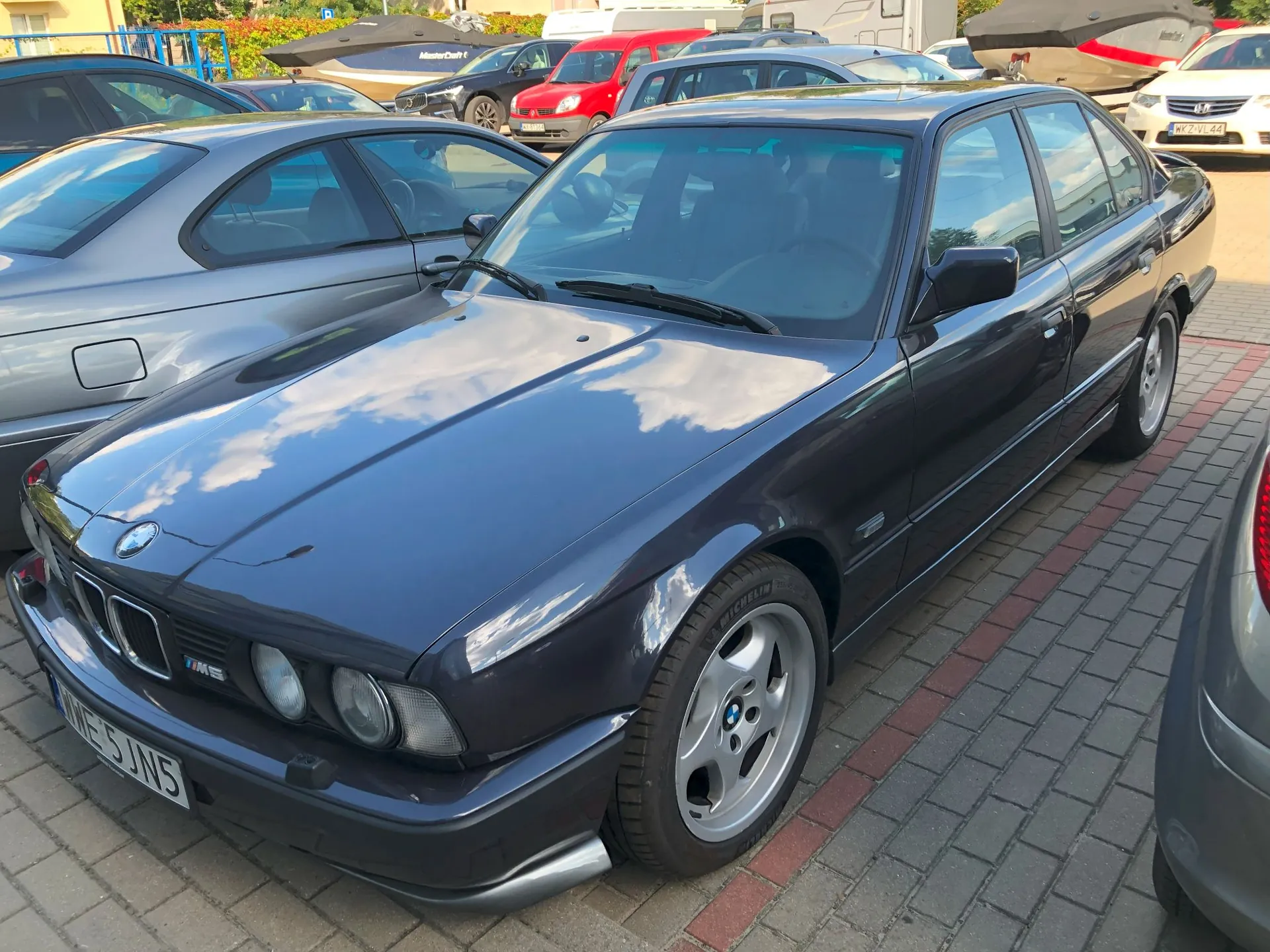 Ciemnogranatowy samochód BMW serii 5 zaparkowany na kostce brukowej. Auto jest widoczne z przodu i z boku, z wyraźnym logo BMW na masce i felgami aluminiowymi. W tle widoczne są inne zaparkowane samochody w różnych kolorach oraz łodzie na przyczepach.