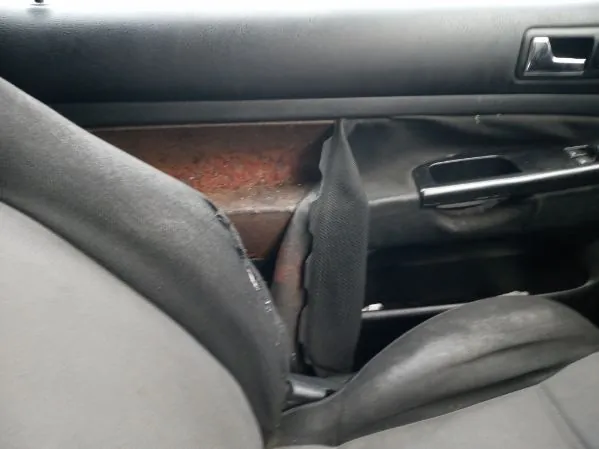 Wnętrze samochodu z widocznym szarym siedzeniem, częścią drzwi z rdzą na metalowej powierzchni, czarną klamką drzwiową, mechanizmem sterującym oknem oraz pasem bezpieczeństwa. Brak czytelnego tekstu.