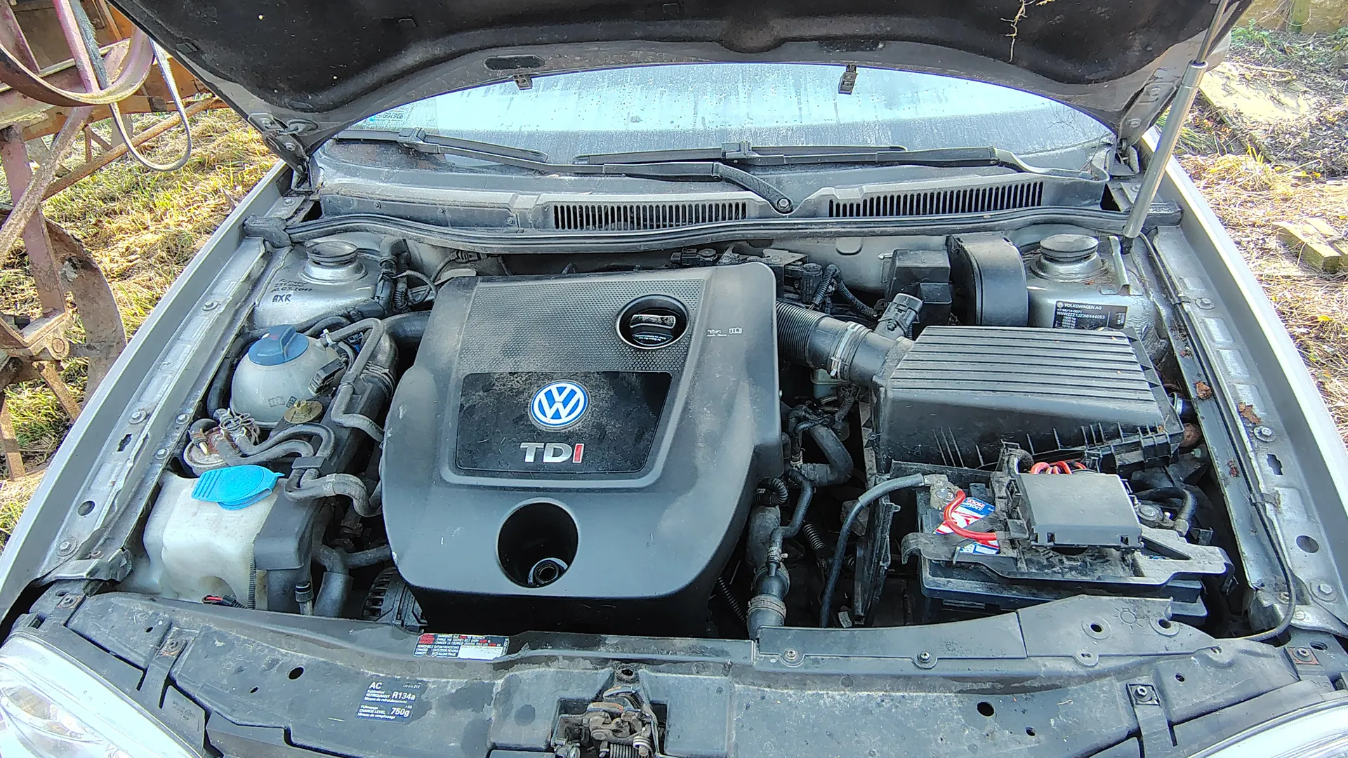 Otwarty kapota samochodu z widocznym silnikiem marki Volkswagen TDI. Na pokrywie silnika widoczne są logo VW oraz napis "TDI". W komorze silnika znajdują się elementy takie jak zbiornik chłodniczy z niebieską pokrywką, przewody, filtr powietrza oraz inne części mechaniczne. Samochód stoi na zewnątrz, na trawie, z częścią metalowej konstrukcji po lewej stronie kadru.
