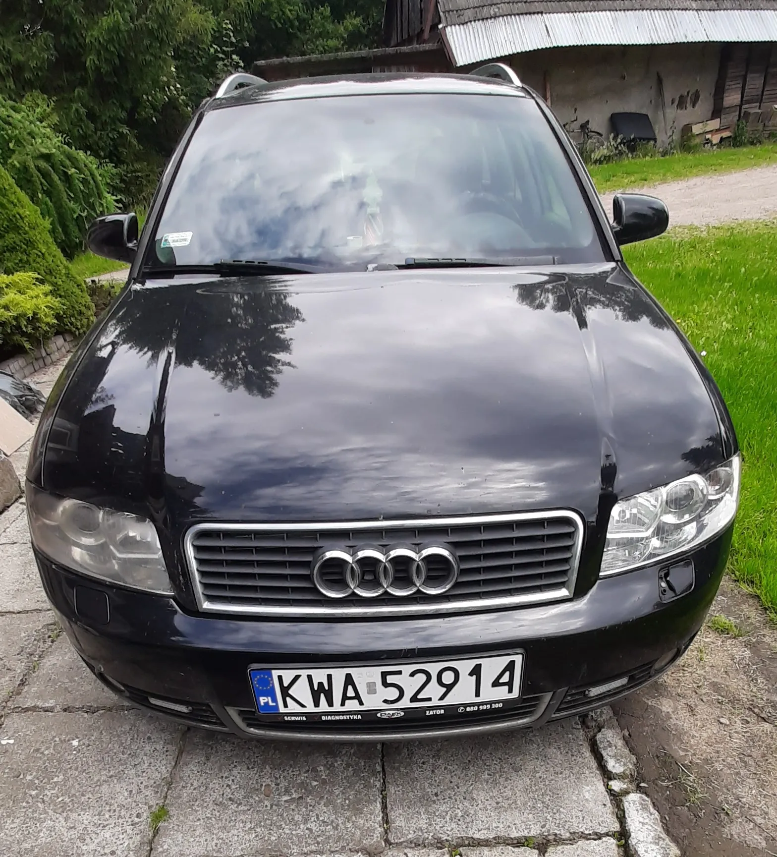 Czarny samochód marki Audi zaparkowany na betonowych płytach. Widoczny jest przód pojazdu z charakterystycznym grillem i logo czterech pierścieni. Samochód posiada polską tablicę rejestracyjną o numerze KWA 52914. W tle widać zieloną roślinność oraz fragment budynku z szarym dachem.