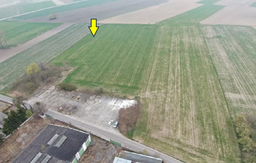 Zdjęcie lotnicze przedstawiające obszar rolniczy z polami w różnych odcieniach zieleni i brązu. W dolnej części obrazu widoczny jest betonowy plac z zaparkowanymi dwoma białymi samochodami oraz fragmenty budynków przemysłowych z ciemnymi dachami. Na górnej części zdjęcia znajduje się żółta strzałka skierowana w dół, wskazująca na zielone pole uprawne.