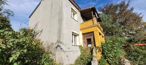 Dwupiętrowy budynek mieszkalny z szarą elewacją i żółtą częścią z balkonem na prawej stronie. Widoczne są dwa okna na szarej ścianie oraz drzwi z metalową kratą na żółtej części. Budynek otoczony jest zielonymi krzewami i drzewami, na tle błękitnego nieba z nielicznymi chmurami. Elewacja budynku wykazuje ślady starzenia się, w tym plamy i uszkodzenia na szarej ścianie.