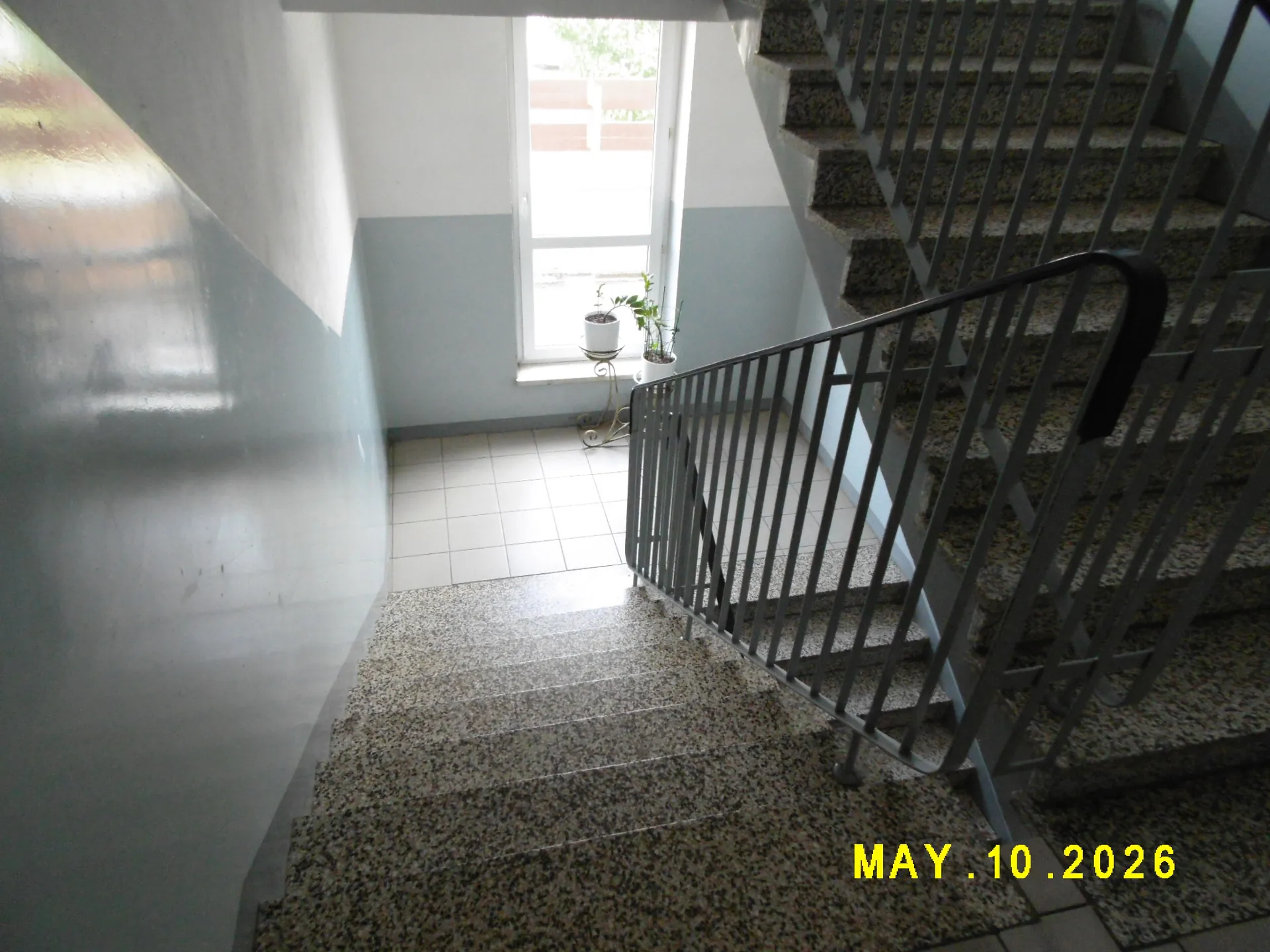 Widok na klatkę schodową z granitowymi stopniami. Po prawej stronie znajdują się schody prowadzące w górę z metalową balustradą. W głębi znajduje się jasny korytarz z białymi kafelkami na podłodze i oknem, przy którym stoi mała roślina w doniczce na białym stoliku. W prawym dolnym rogu zdjęcia widnieje żółty napis z datą: MAY .10 .2026