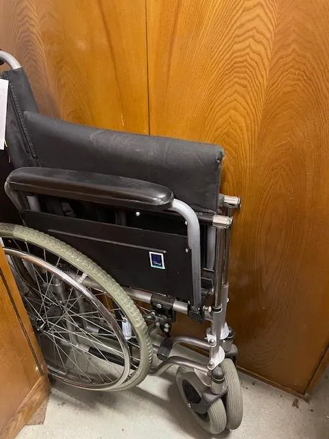 Wózek inwalidzki z czarnym siedziskiem i oparciem, metalową ramą, dużymi kołami z obręczami do napędu i małymi przodowymi kółkami. Na tle drewnianych paneli o pionowym usłojeniu. Na ramie wózka widoczny mały niebieski i biały naklejony znaczek. Podłoga wyłożona jasnym materiałem.