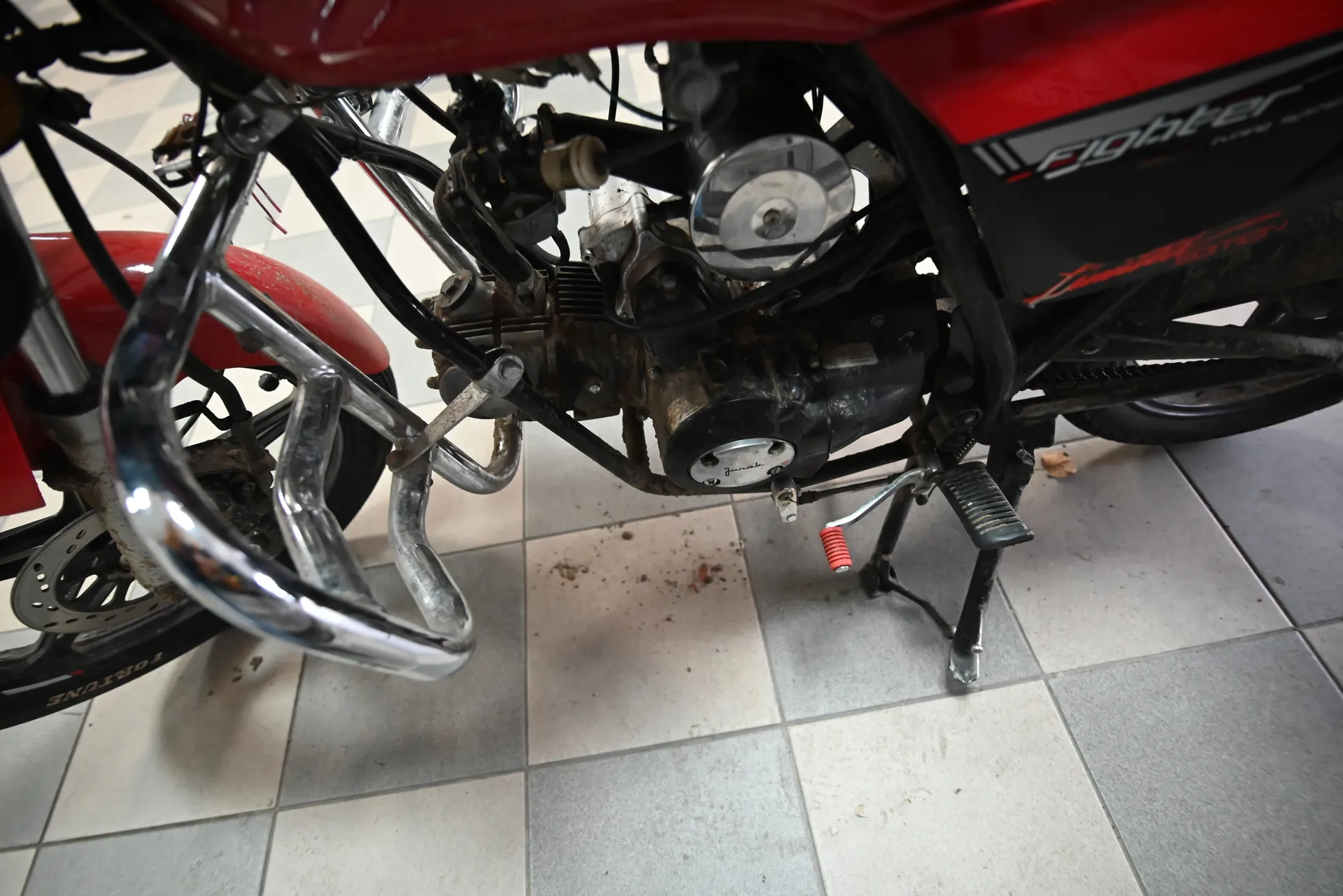 Czerwony motocykl z widocznym silnikiem, na boku napis "Fighter" oraz "FORTUNE" na oponie. Chromowane rury wydechowe, podnóżek z czerwoną sprężyną, brudny silnik i podłoga z szarymi płytkami z resztkami brudu. Motocykl oparty na stojaku.