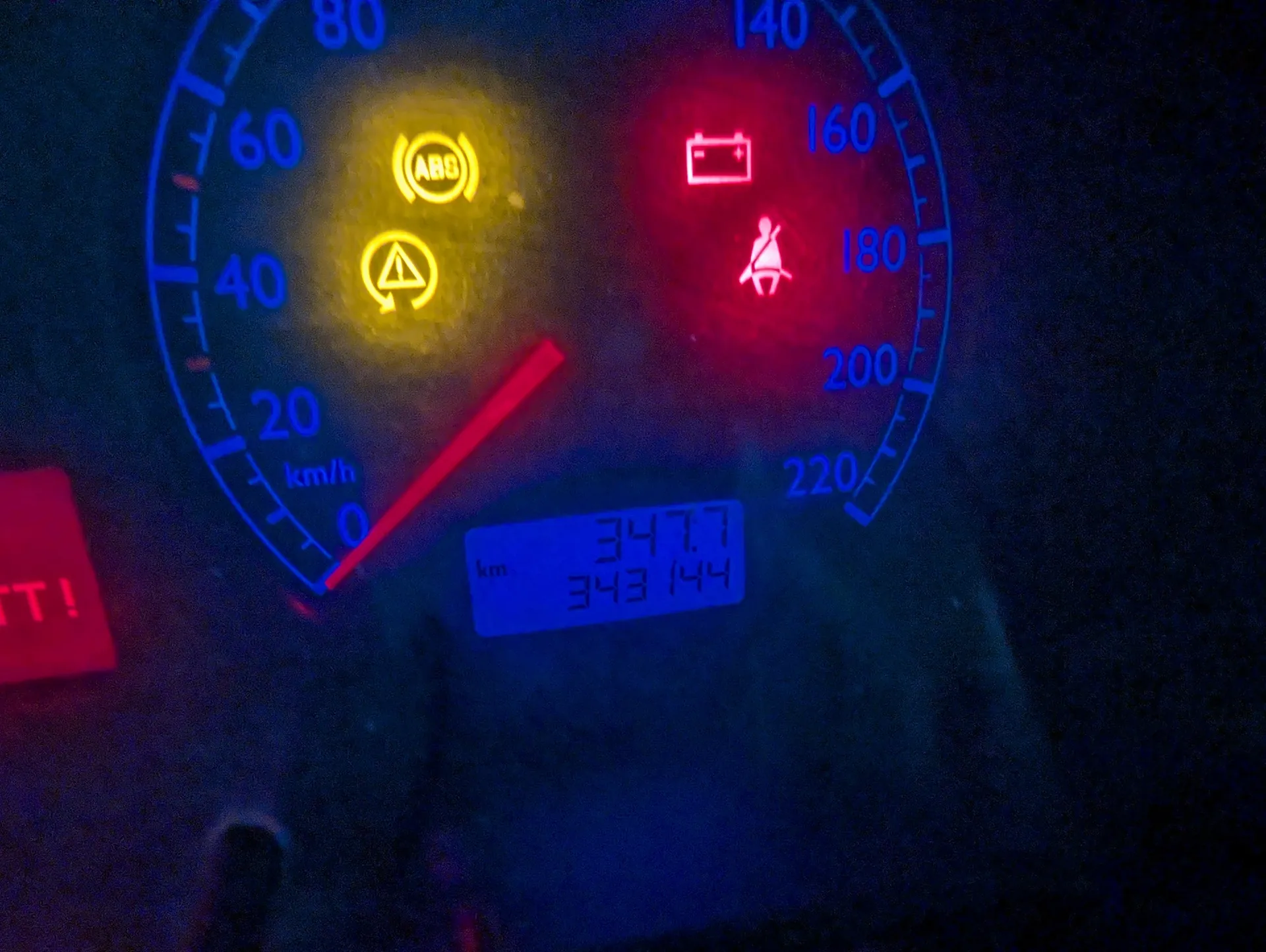 Zbliżenie na podświetlony licznik prędkości w samochodzie w ciemności. Tarcza prędkościomierza ma niebieskie oznaczenia i skalę od 0 do 220 km/h. Na tarczy widoczne są żółte kontrolki ABS oraz ostrzegawcza (trójkąt z wykrzyknikiem) oraz czerwone kontrolki akumulatora i pasów bezpieczeństwa. Poniżej znajduje się niebieski wyświetlacz cyfrowy z liczbami 347.7 oraz 343 144. Po lewej stronie widoczny jest fragment czerwonego wyświetlacza z tekstem 'TT!'
