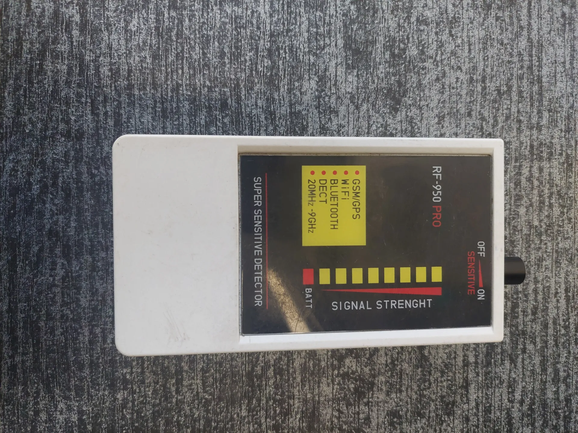 Biały detektor sygnału o prostokątnym kształcie z czarnym wyświetlaczem. Na wyświetlaczu widoczne są napisy: "RF-950 PRO", "SIGNAL STRENGTH" z czerwoną linią i żółtymi kwadratami wskazującymi siłę sygnału, "BATT" z czerwonym kwadratem oraz "SUPER SENSITIVE DETECTOR". W żółtym polu znajdują się napisy: "GSM/GPS", "WiFi", "BLUETOOTH", "DECT", "20MHz-9GHz", każdy z czerwoną kropką. Po lewej stronie wyświetlacza przełącznik z pozycjami "OFF" i "ON" oraz napisem "SENSITIVE". Tło stanowi szara, teksturowana powierzchnia.