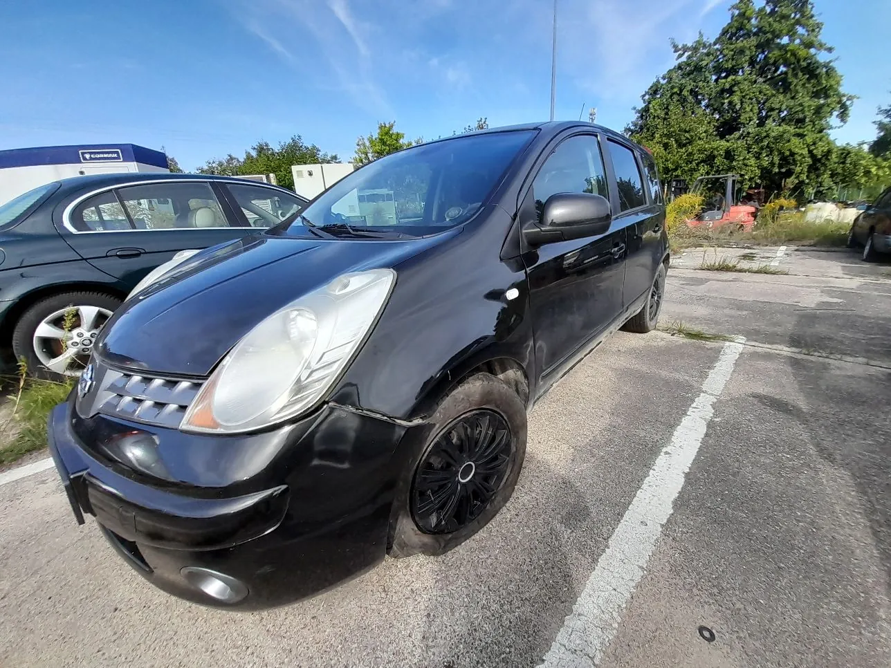 Czarny samochód osobowy typu minivan zaparkowany na betonowym placu. Pojazd ma widoczne uszkodzenia karoserii w przedniej części, szczególnie w okolicach lewego błotnika i zderzaka. W tle widoczny jest inny, ciemny samochód, zielone drzewa oraz fragment białego budynku pod jasnoniebieskim niebem.