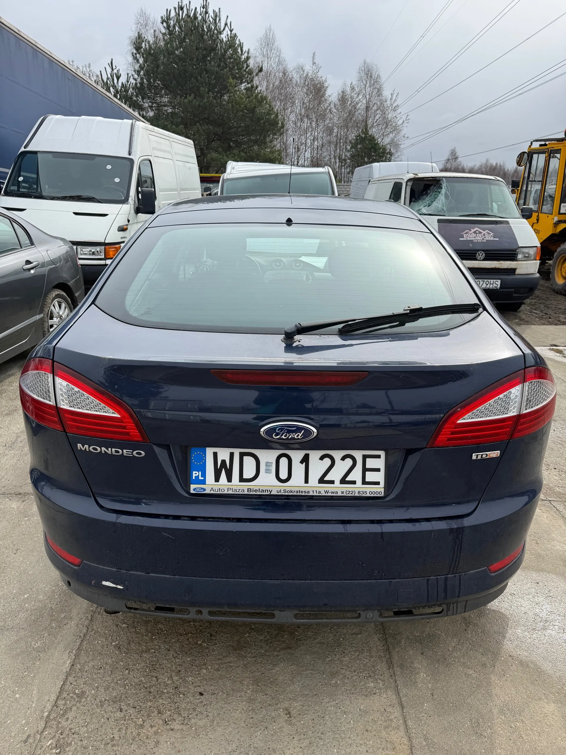 Z tyłu widok ciemnoniebieskiego samochodu Ford Mondeo z tablicą rejestracyjną WD 0122E. Na tablicy widoczny napis PL oraz informacja o salonie Auto Plaza Bielany ul. Sokratesa 11a, W-wa (22) 835 0000. W tle stoją inne pojazdy: białe furgony, szary samochód osobowy i żółty pojazd budowlany. Tło stanowią drzewa, linie energetyczne i pochmurne niebo. Samochód stoi na betonowej powierzchni.