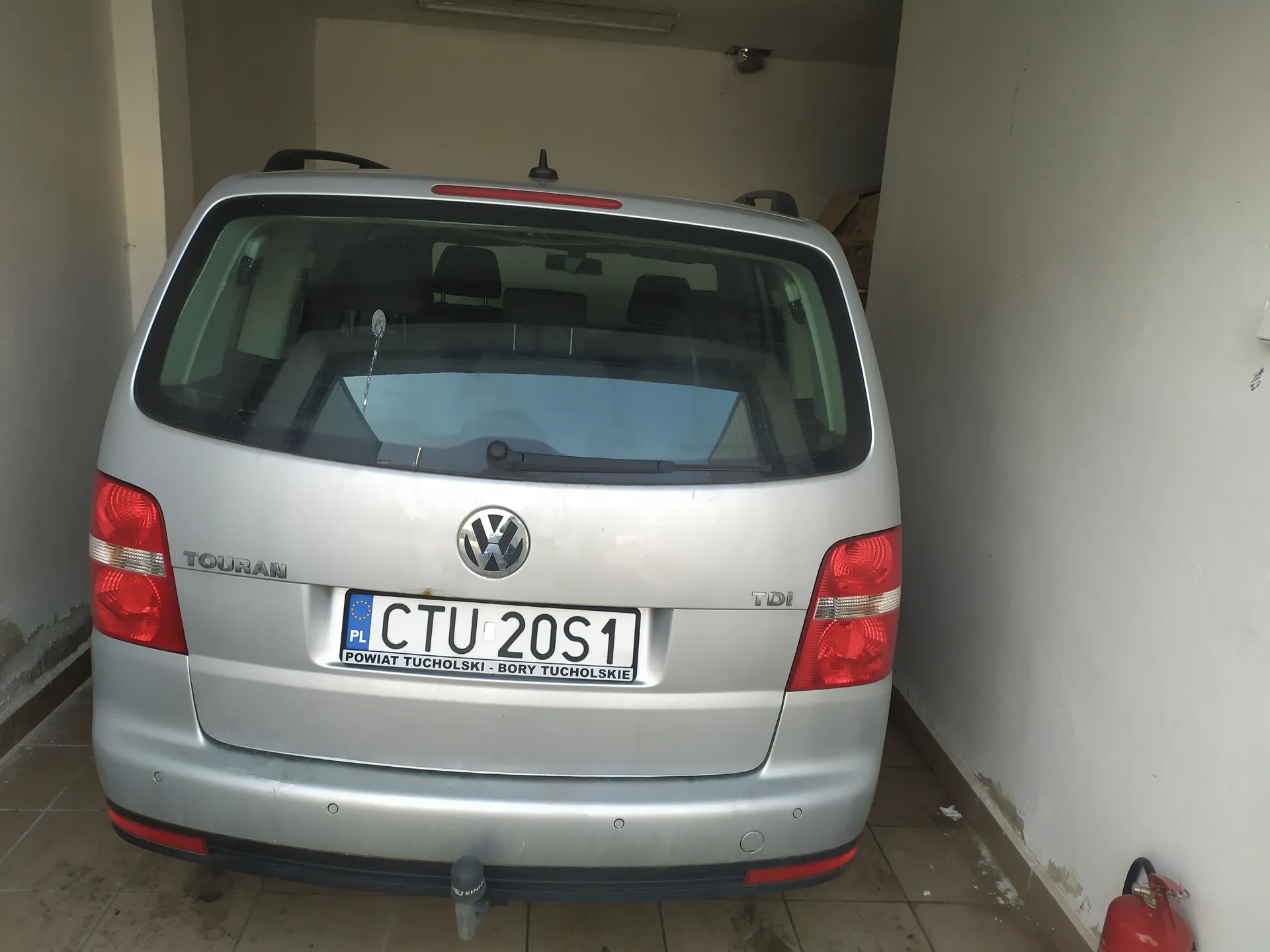 Tył srebrnego samochodu Volkswagen Touran z widocznym logo VW, napisem TOURAN po lewej stronie i TDI po prawej. Tablica rejestracyjna z białym tłem i czarnym tekstem: CTU 20S1, poniżej napis POWIAT TUCHOLSKI - BORY TUCHOLSKIE oraz oznaczenie PL z niebieskim pasem i żółtymi gwiazdkami. Samochód zaparkowany w garażu z białymi ścianami i podłogą z kafli. Po prawej stronie widoczny czerwony gaśnicz. Czerwone światła tylnego oświetlenia i czarna osłona bagażnika.