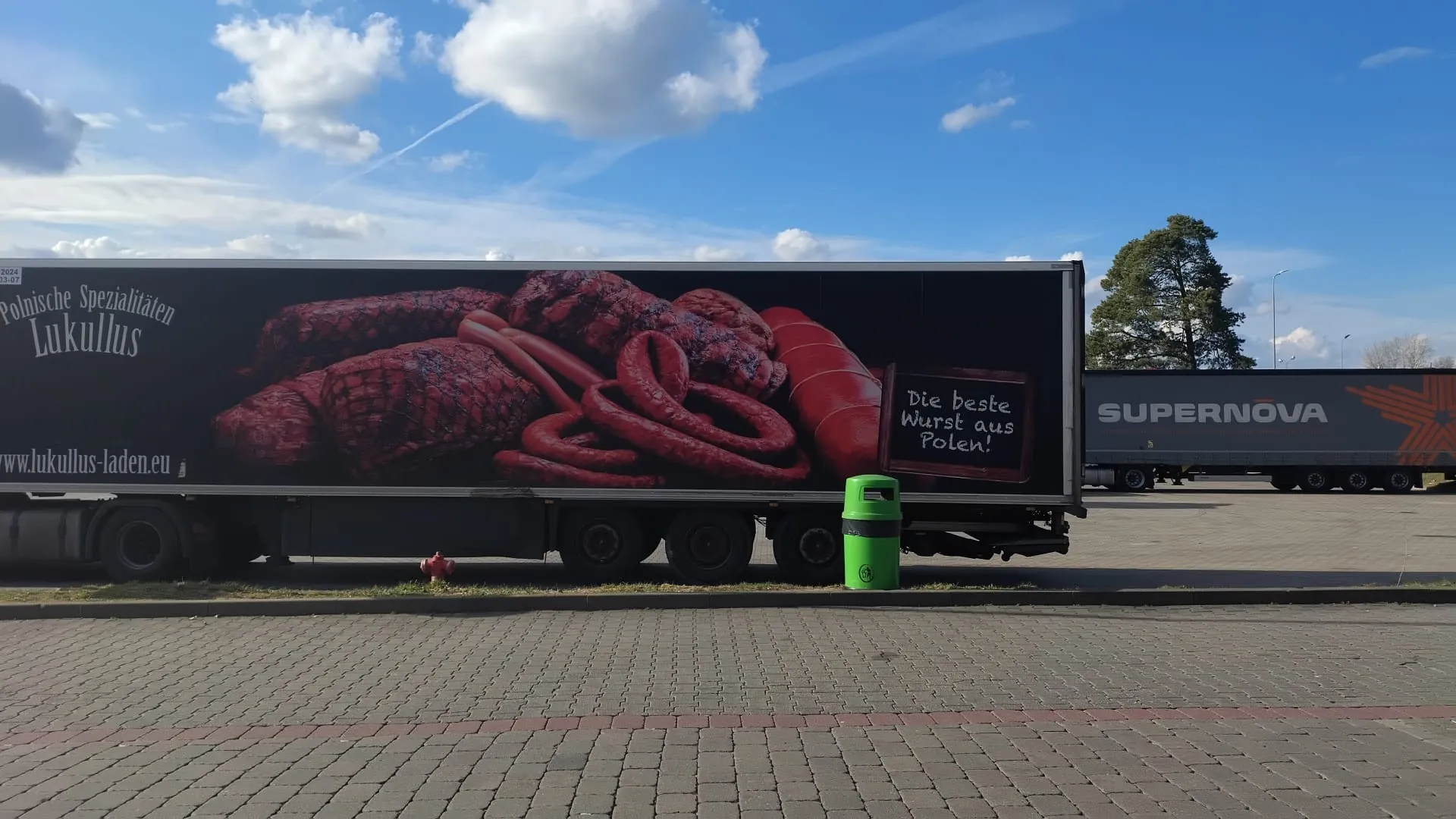 Reklama na boku ciężarówki przedstawiająca różne rodzaje kiełbas i mięsa. Tekst w języku niemieckim: 'Polnische Spezialitäten Lukullus', 'Die beste Wurst aus Polen!' oraz adres strony internetowej 'www.lukullus-laden.eu'. W tle druga ciężarówka z napisem 'SUPERNOVA', zielony kosz na śmieci, czerwony hydrant, brukowana droga i niebo z białymi chmurami.